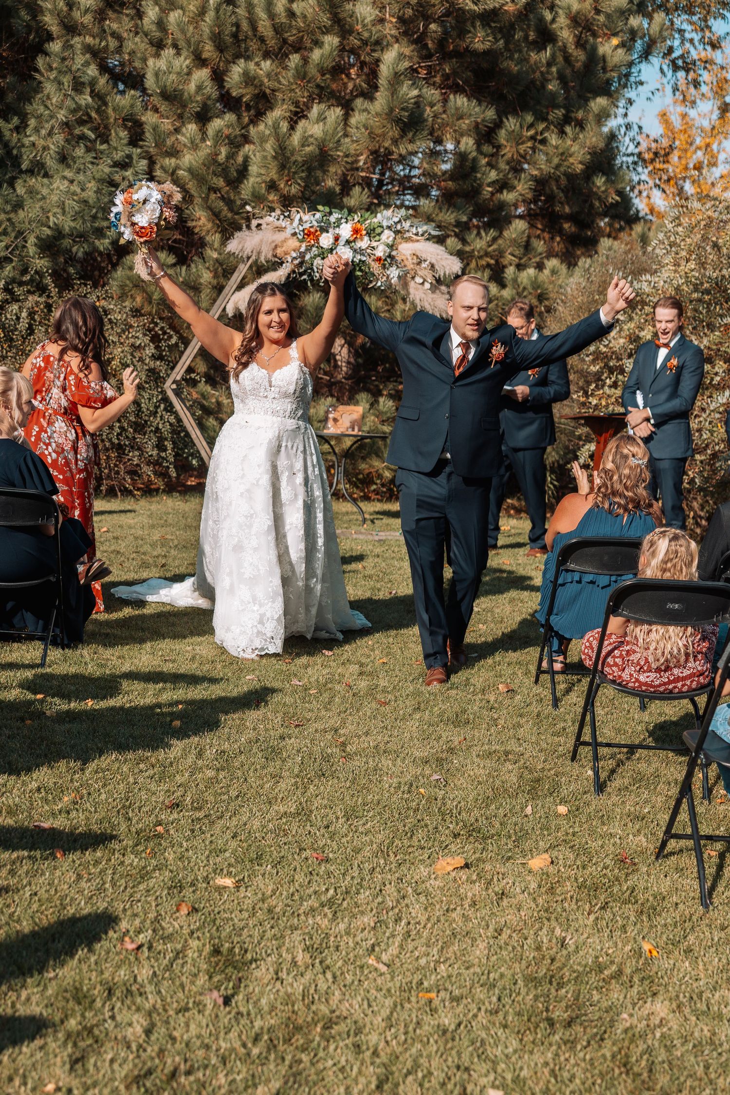Newlyweds celebrate with arms raised while walking down aisle at outdoor ceremony.