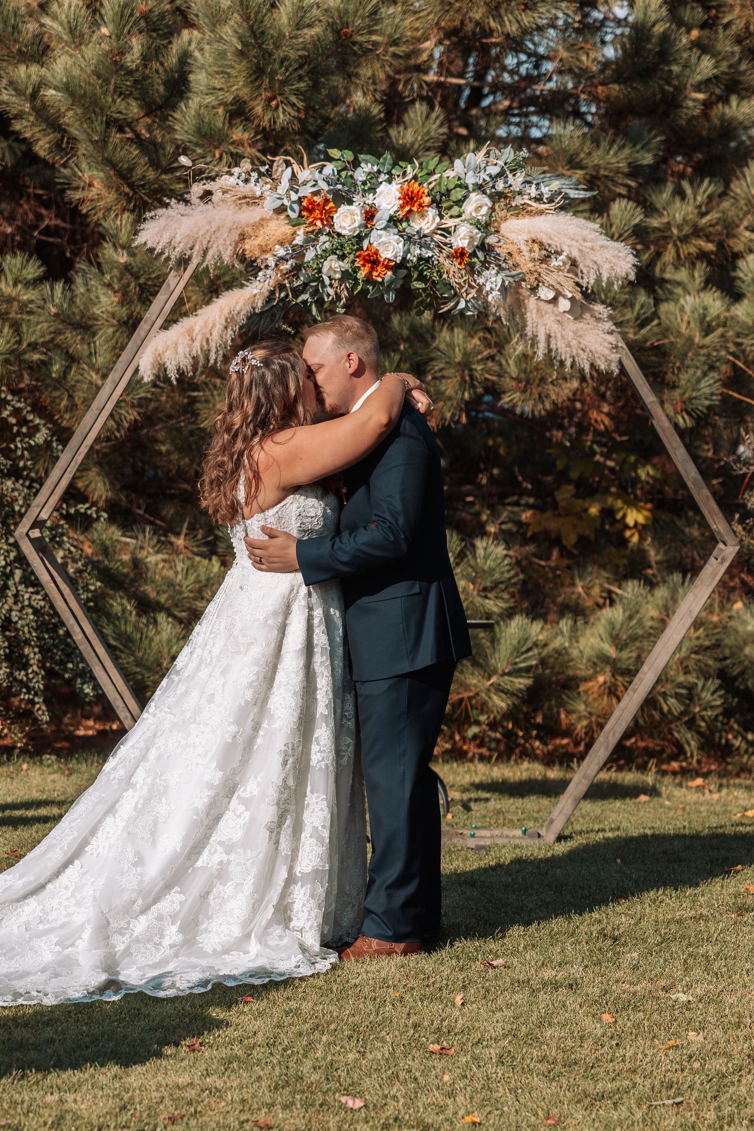 Newlyweds embrace under hexagonal arch decorated with flowers and pampas grass.