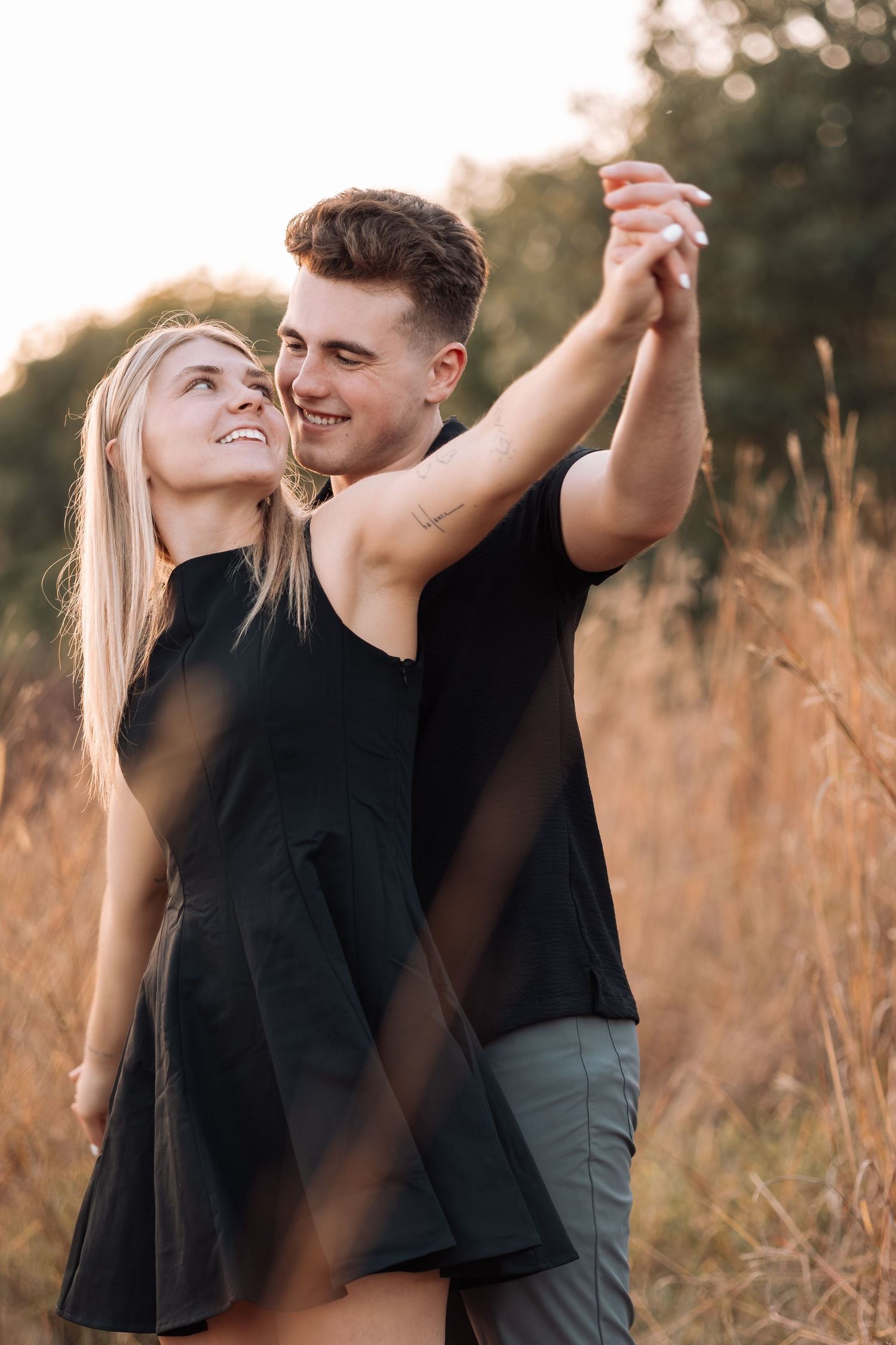 Couple dancing together in a golden field at sunset wearing black outfits.