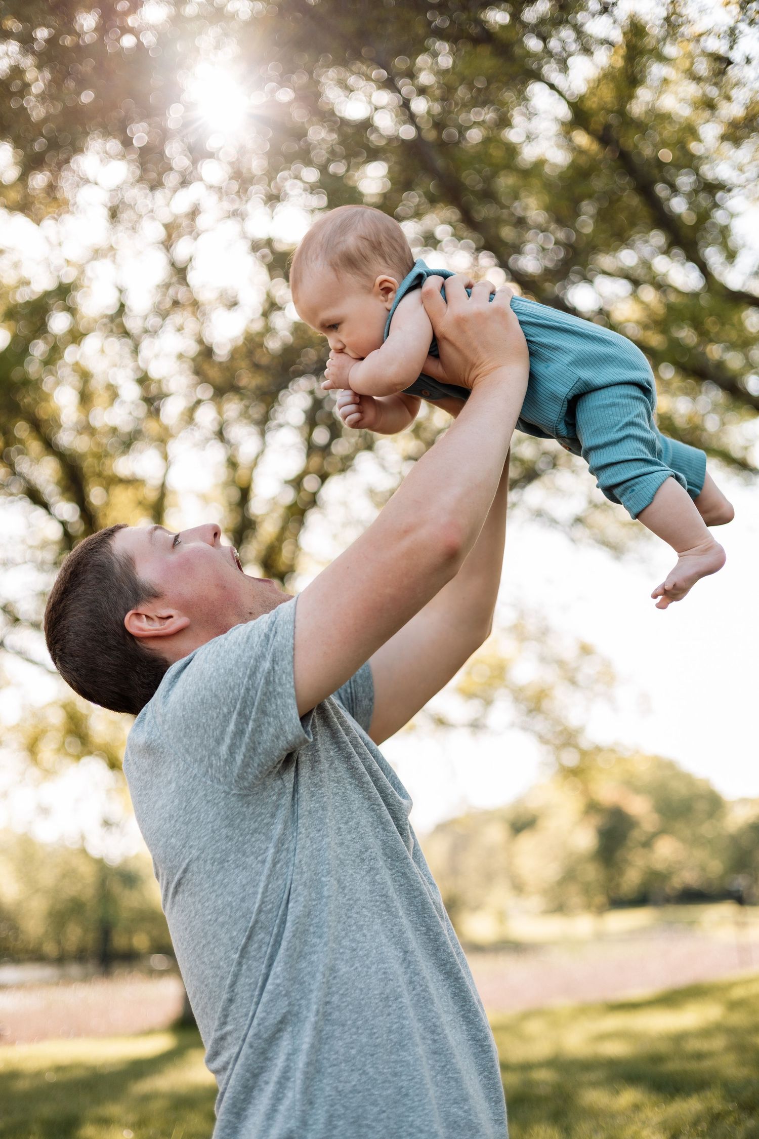 Parent lifts smiling baby in air during outdoor photoshoot in sunlit park.