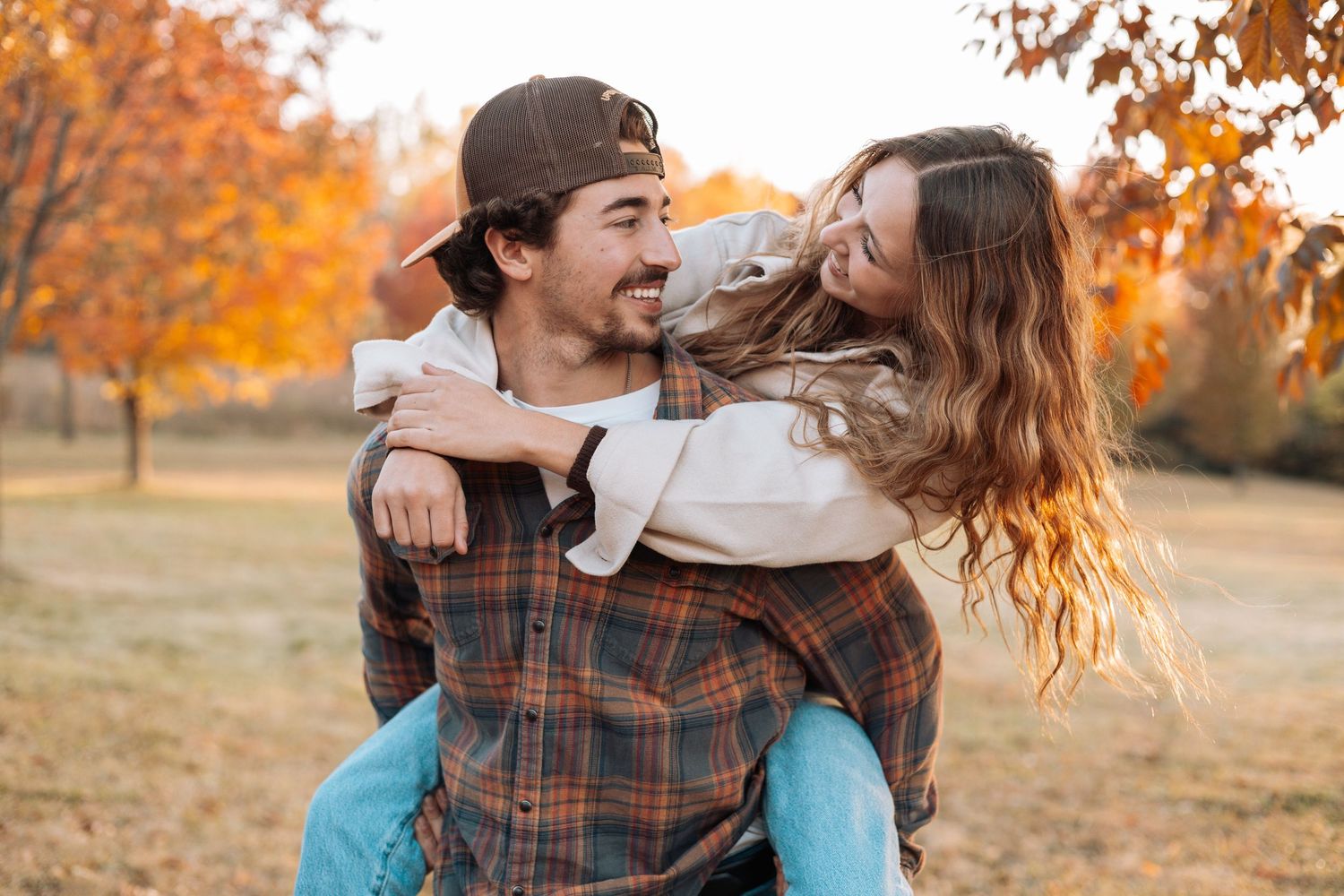 A playful couple shares a piggyback ride in a park with autumn foliage.