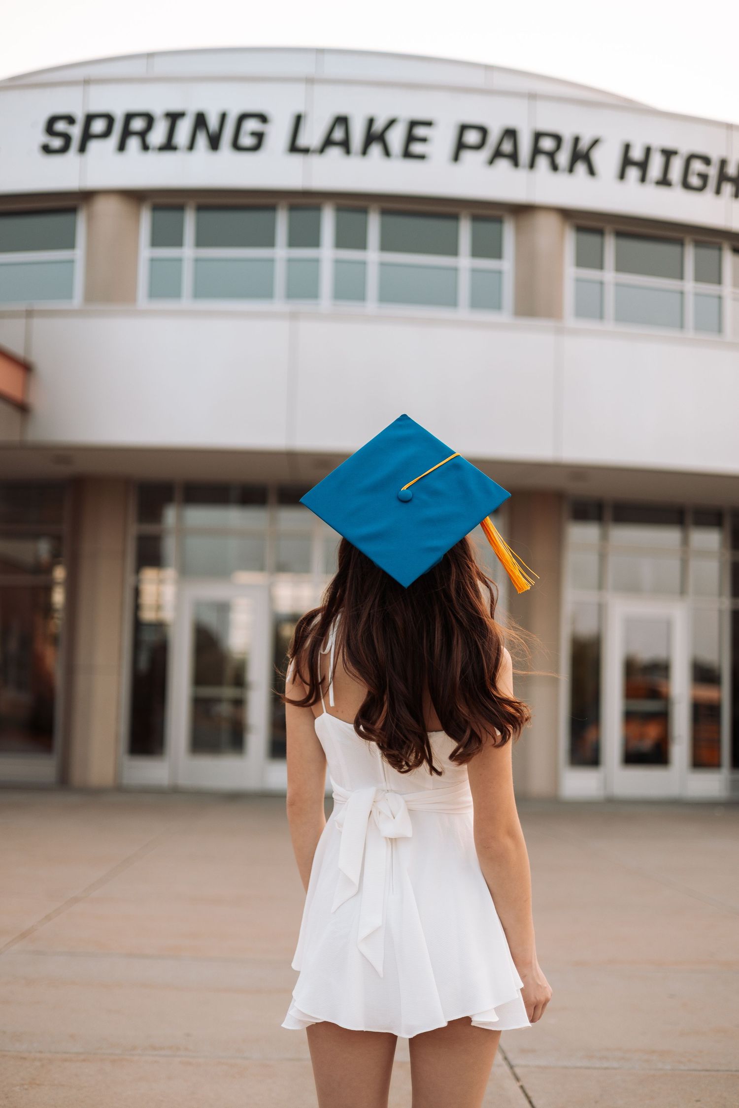 Graduate in white dress faces school building wearing blue graduation cap with Kenzie Ayla Images in the Twin Cities MN area.