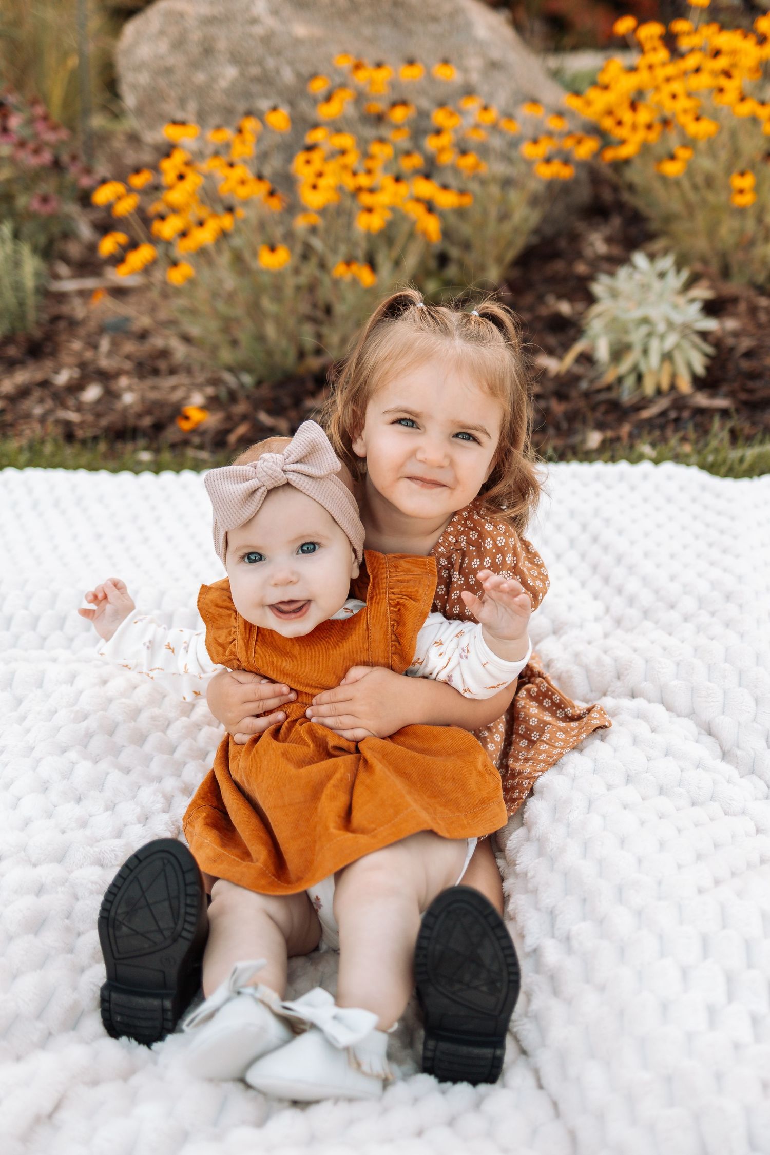 Children in autumn outfits pose on a textured white blanket with bright marigolds blooming behind them.
