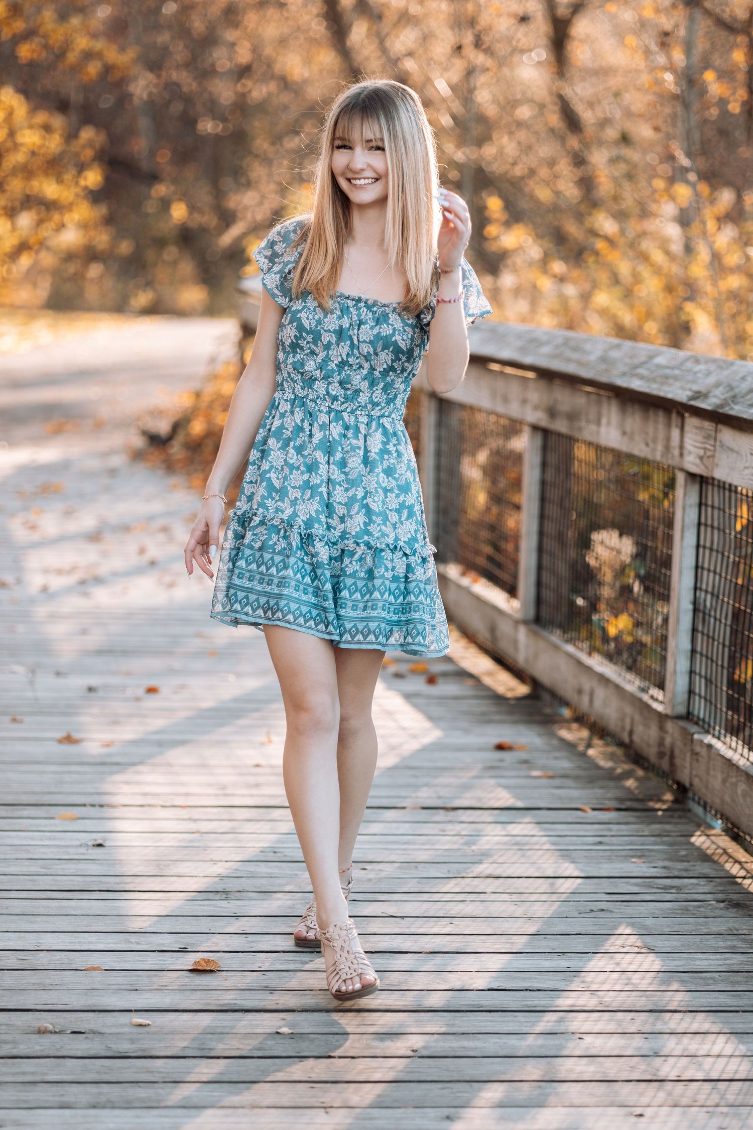 Woman in blue floral dress walks on wooden bridge during sunset with Kenzie Ayla Images in the Twin Cities MN area.