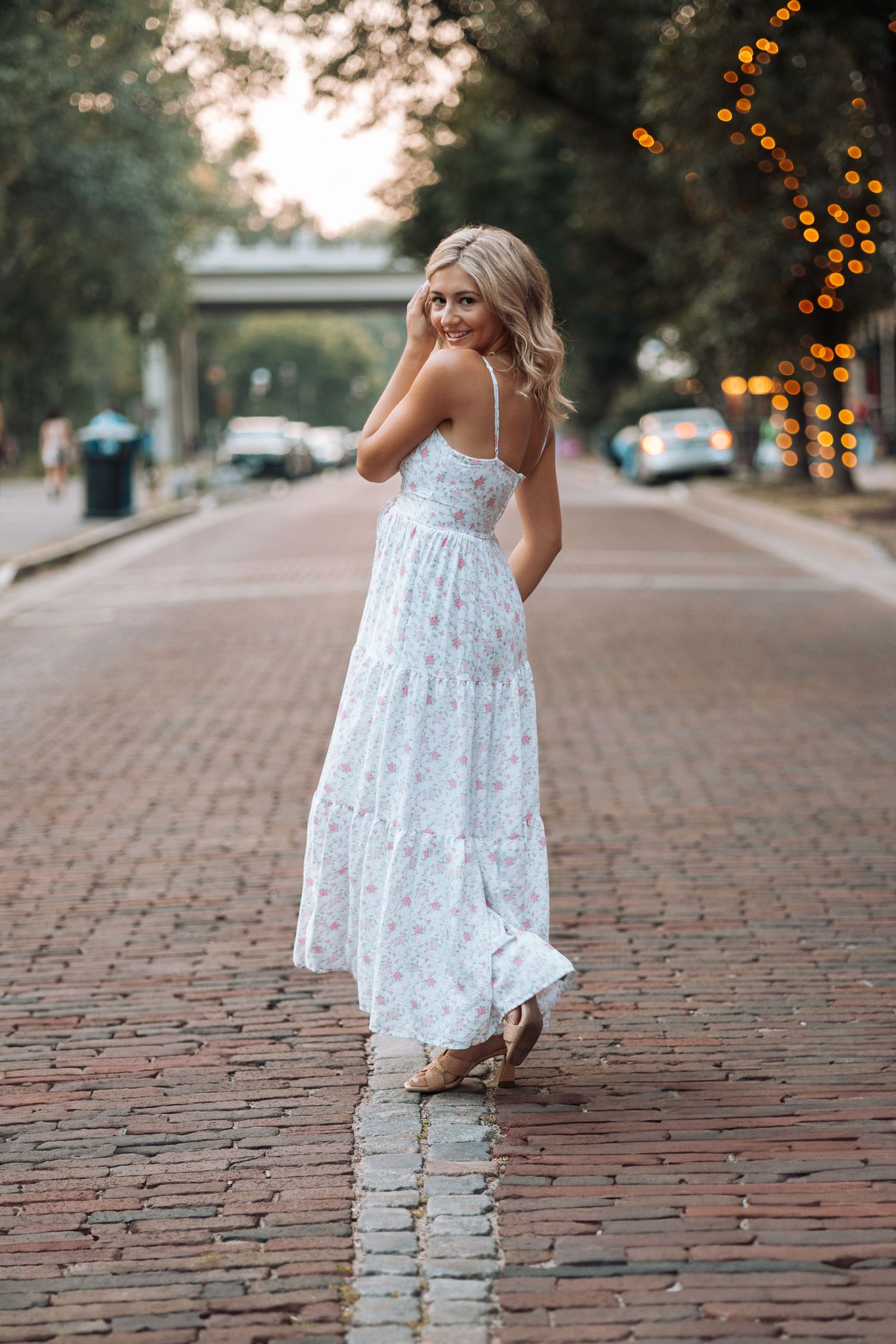 Person in white floral dress poses on brick street with string lights in background with Kenzie Ayla Images in the Twin Cities MN area.