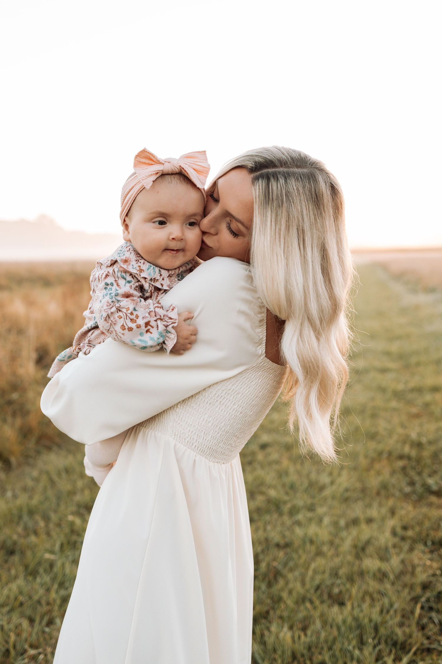 Mother and baby share tender moments in white outfits against a sunset backdrop.
