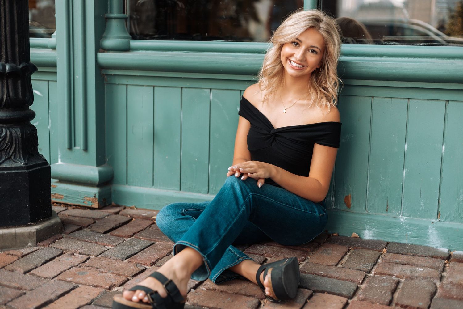 Person in black top and jeans sitting casually against a turquoise wall with Kenzie Ayla Images in the Twin Cities MN area.