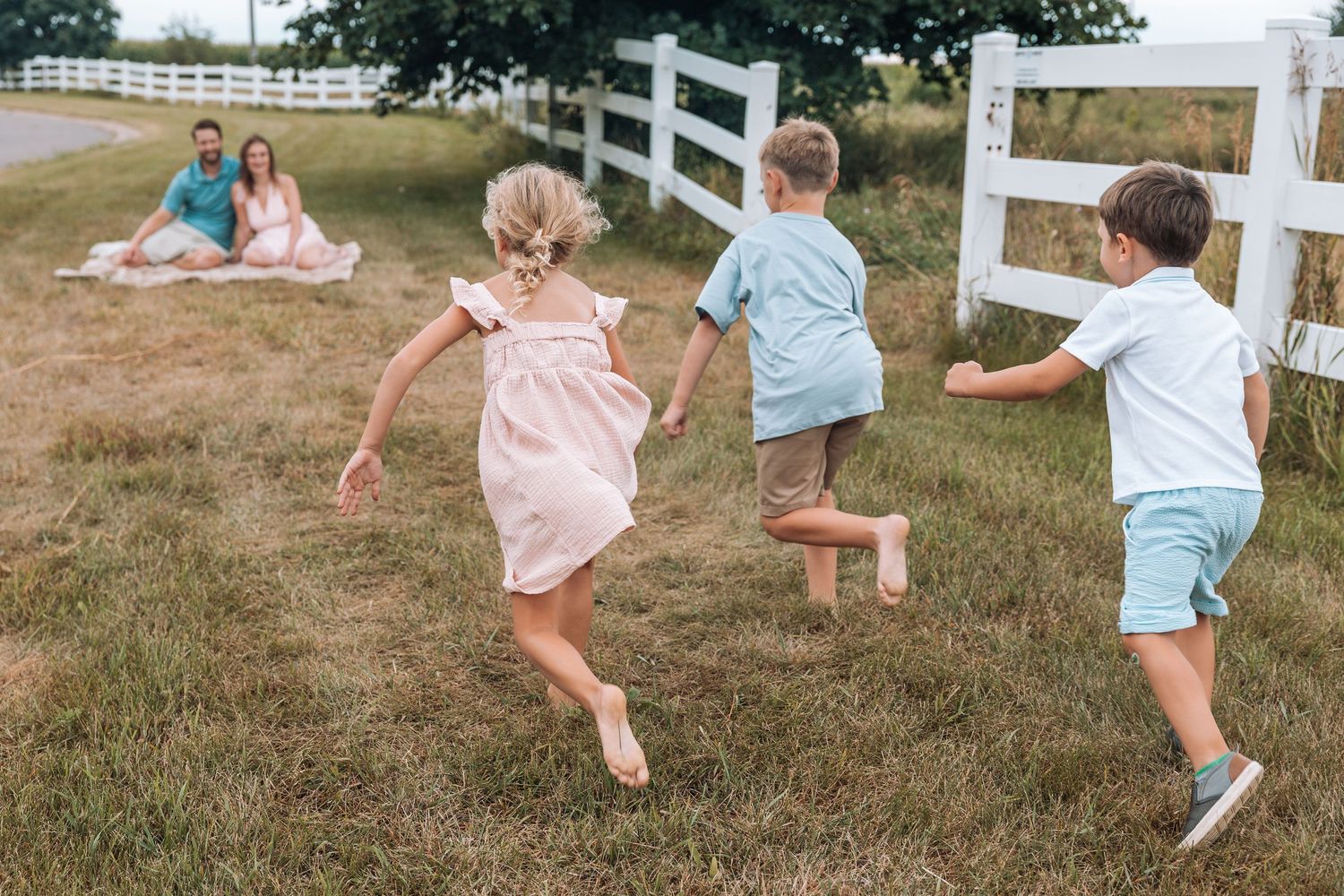 Three children running toward parents sitting on a blanket in a grassy field near a white fence.