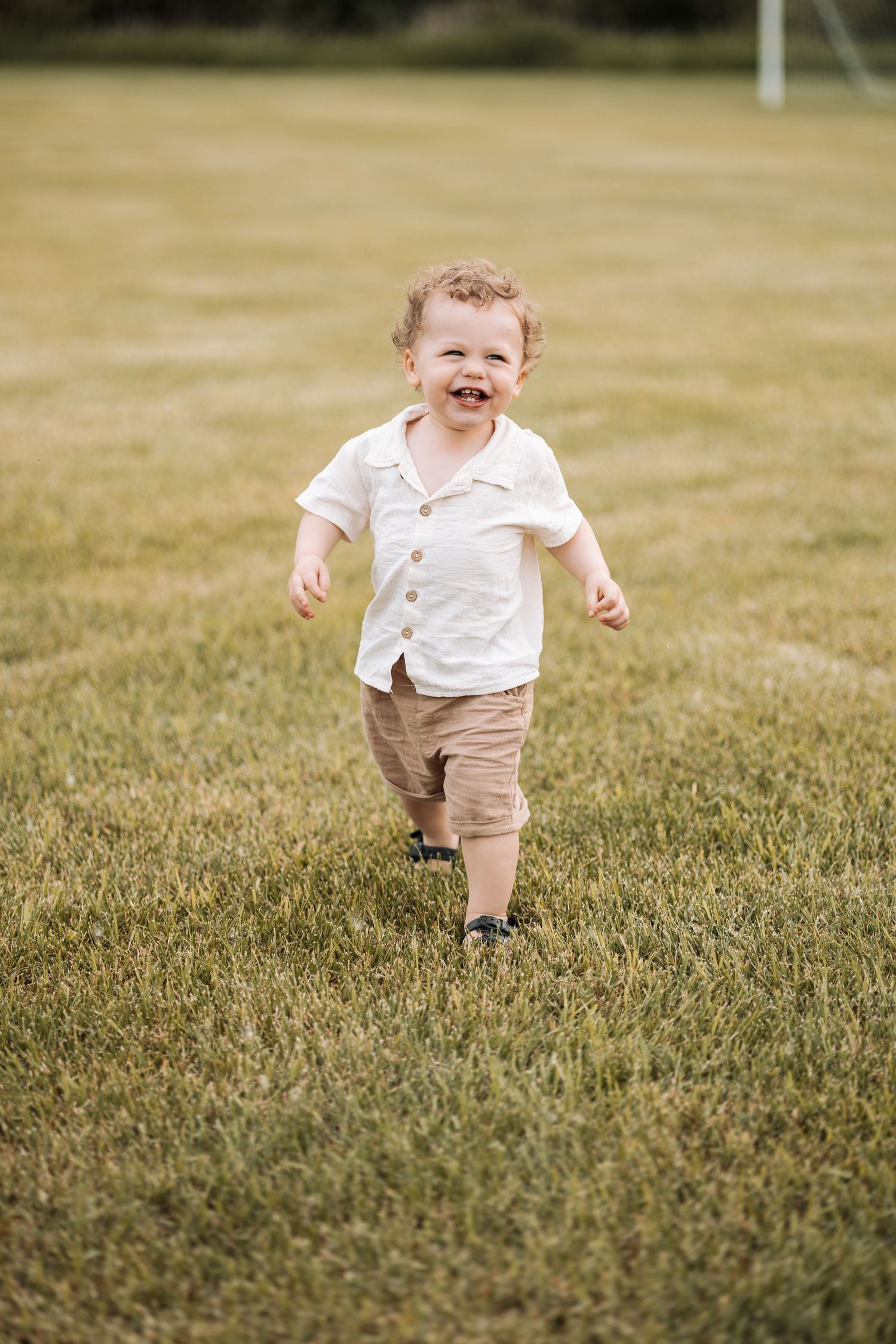 Toddler in white shirt and khaki shorts running happily across green grass field.