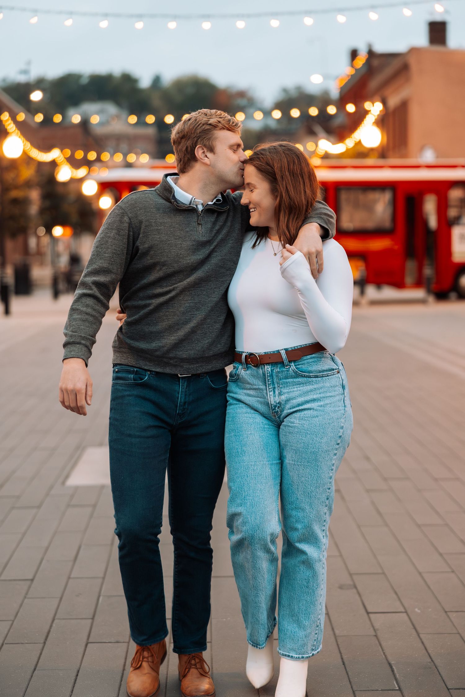 Couple walking together at dusk with string lights and food trucks in background.