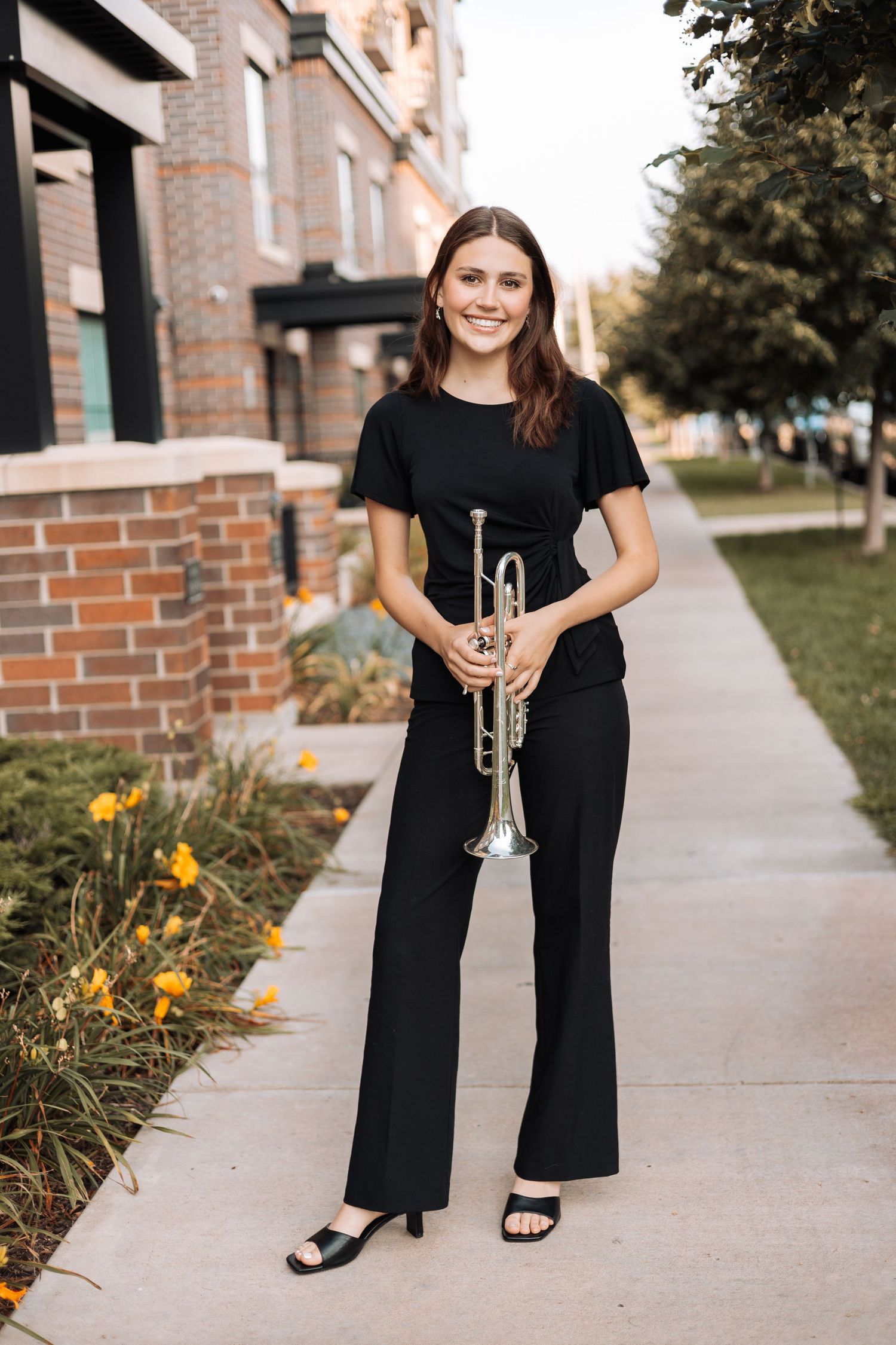 senior in black jumpsuit poses with trumpet on brick sidewalk near yellow flowers with Kenzie Ayla Images in the Twin Cities MN area.