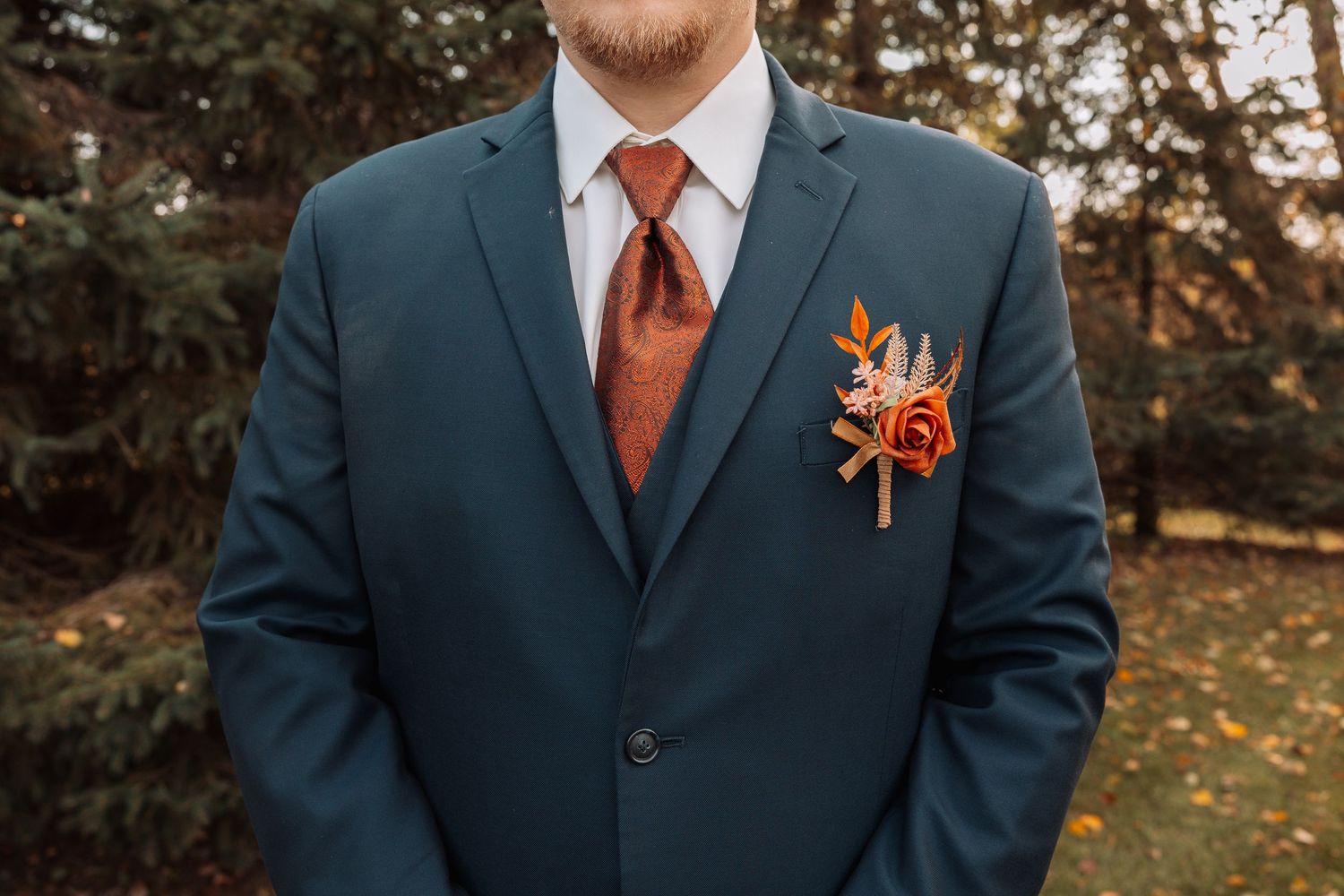 Close-up of navy suit with burnt orange tie and boutonniere against fall foliage.