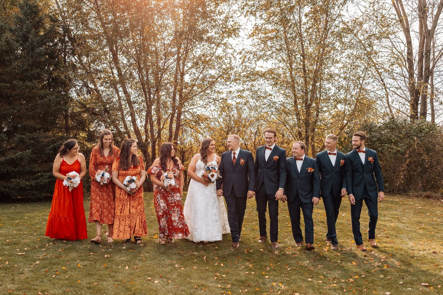 Wedding party lined up outdoors with autumn trees and fallen leaves in background.