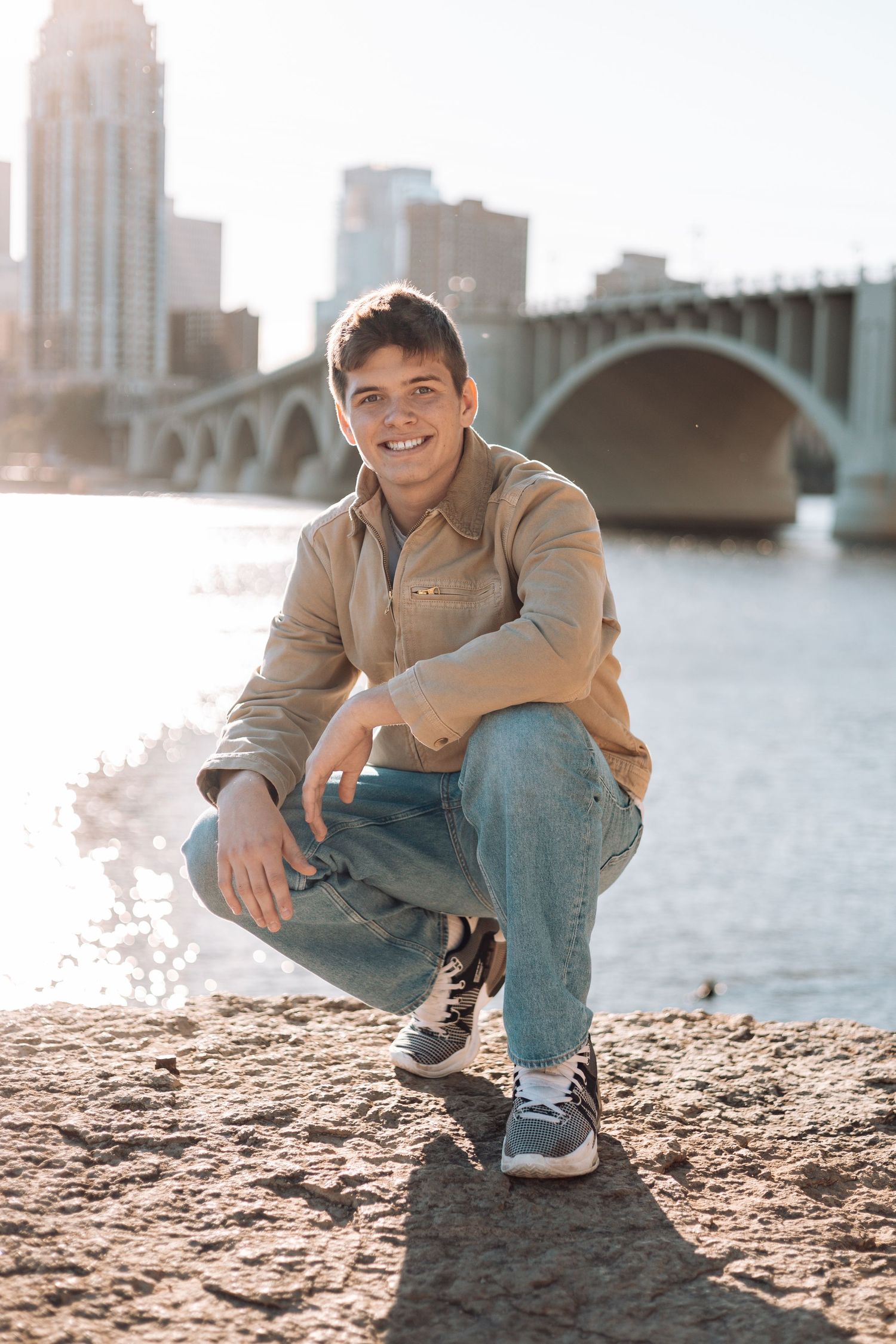 Person in casual attire sits on rocky riverbank with city skyline and bridge in background during golden hour.