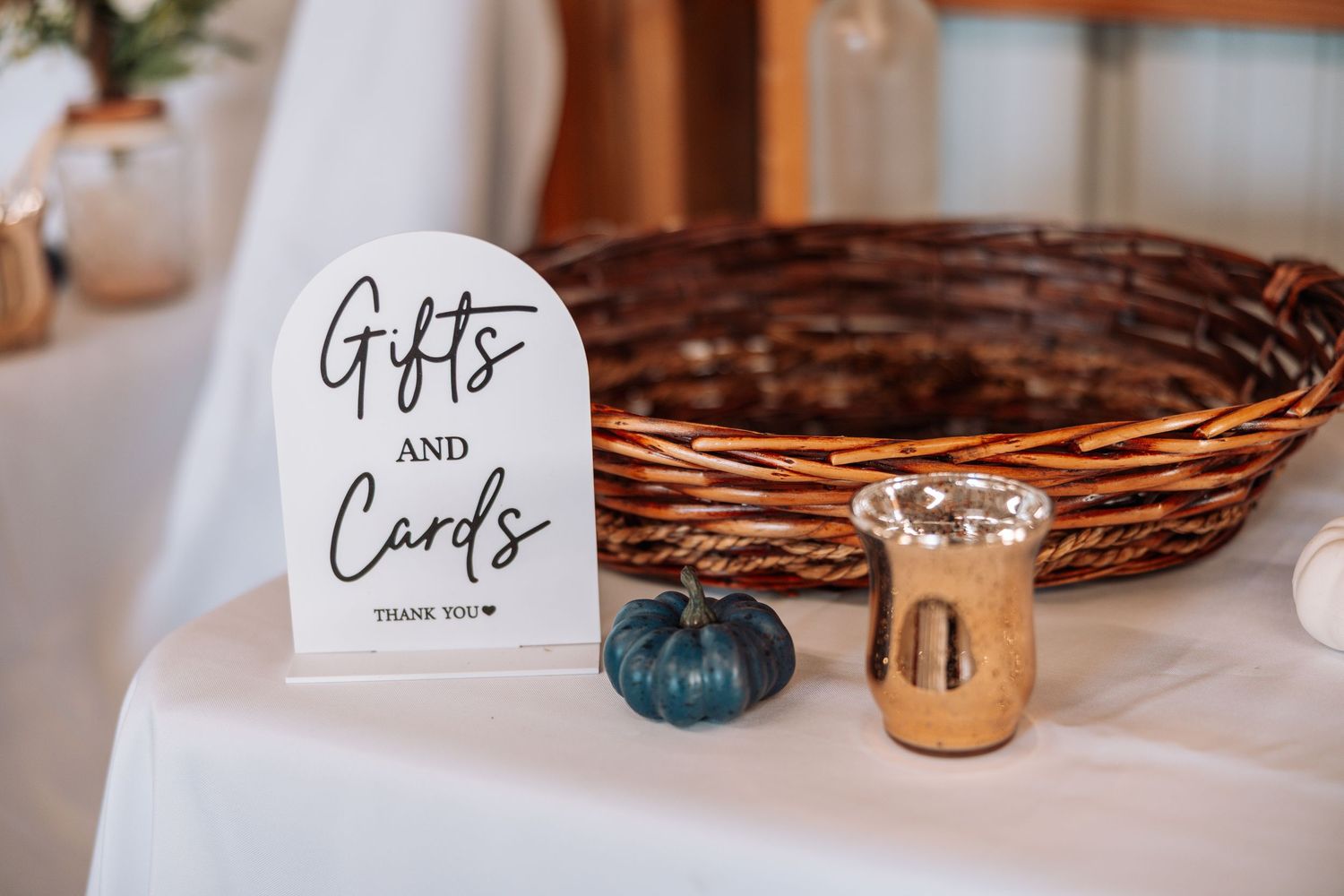 Woven gift basket with 'Gifts and Cards' sign, teal pumpkin, and gold votive holder on white tablecloth.