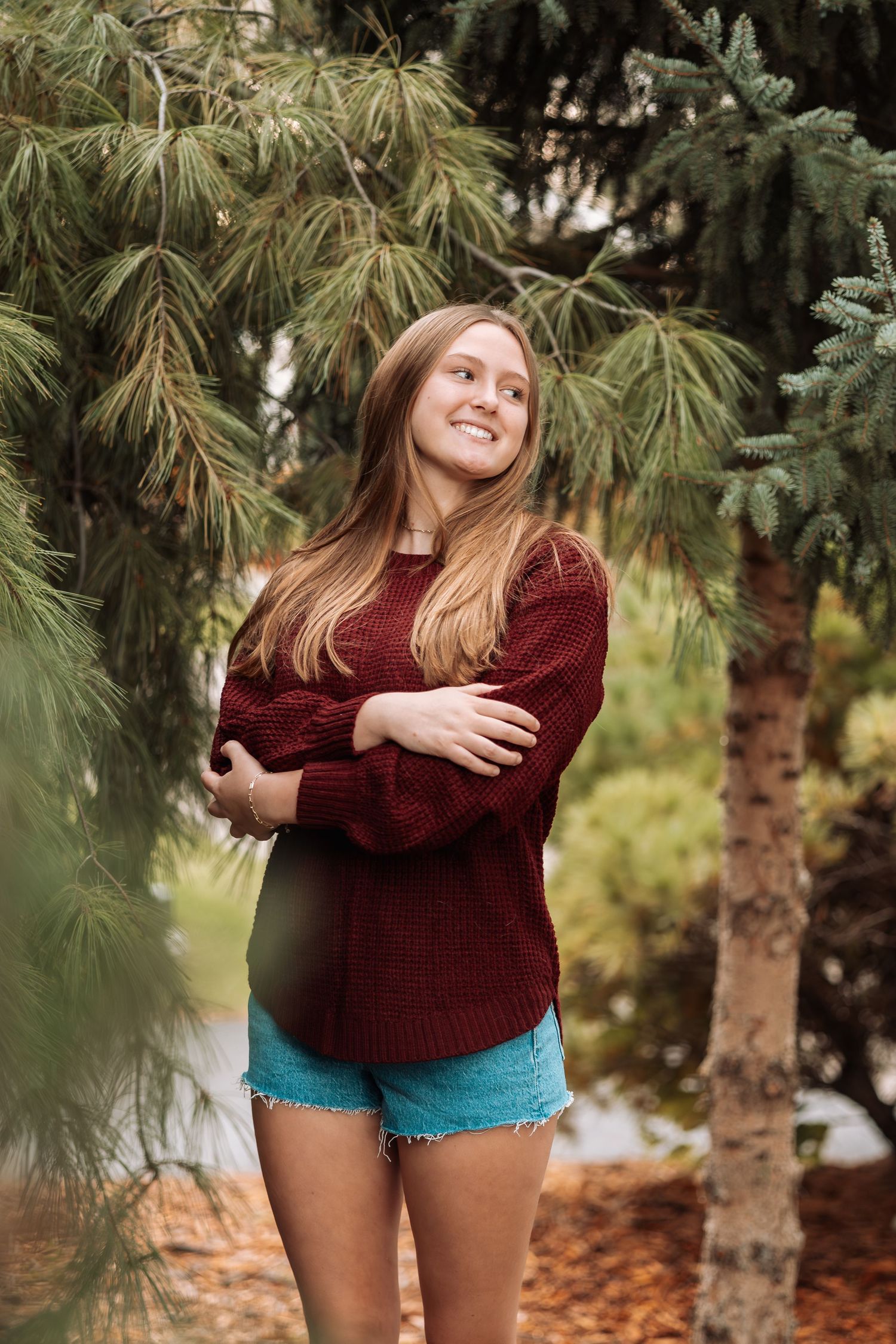 Person in maroon sweater and denim shorts posing near pine trees.