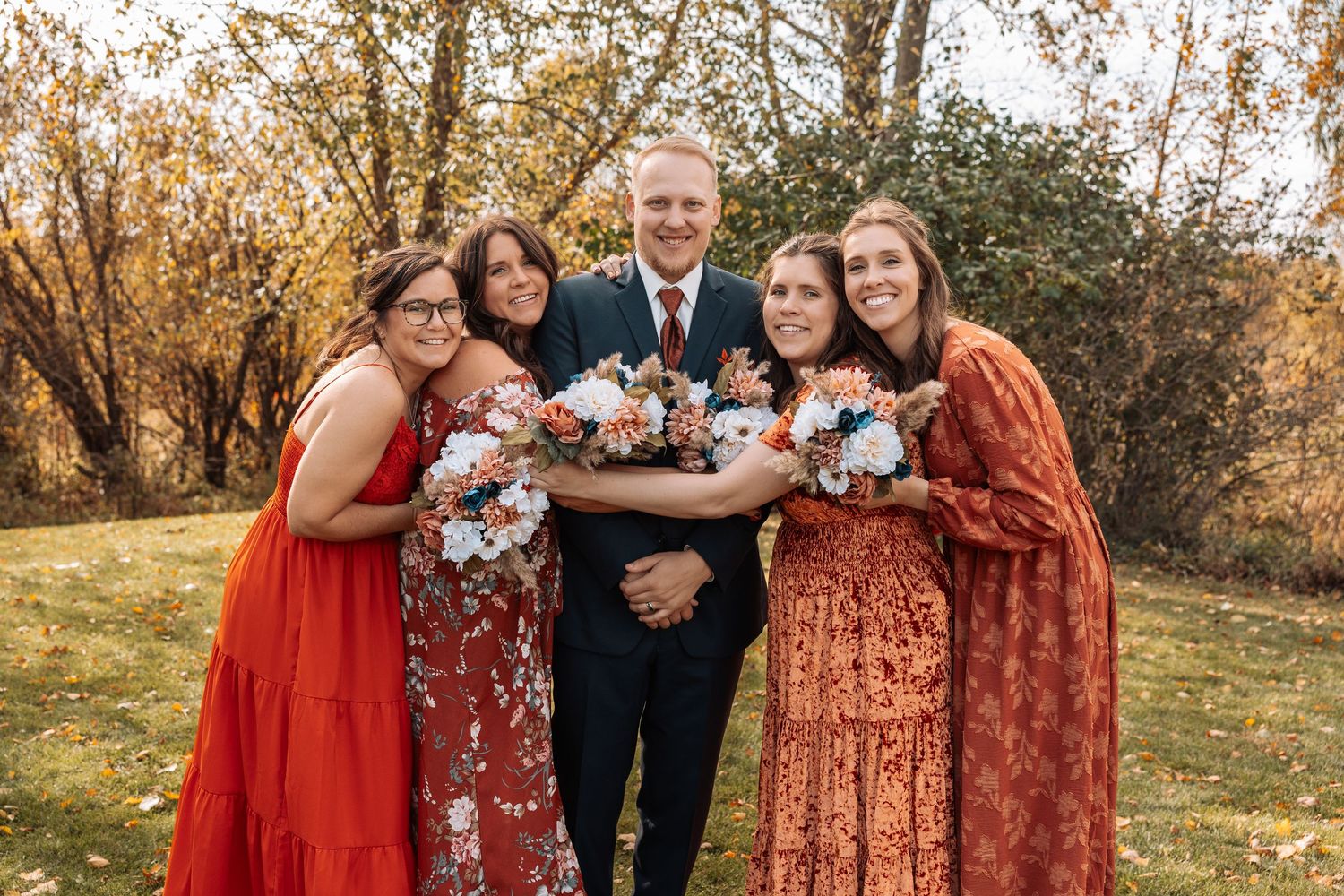 Wedding party in rust and burgundy dresses poses together with bouquets outdoors.