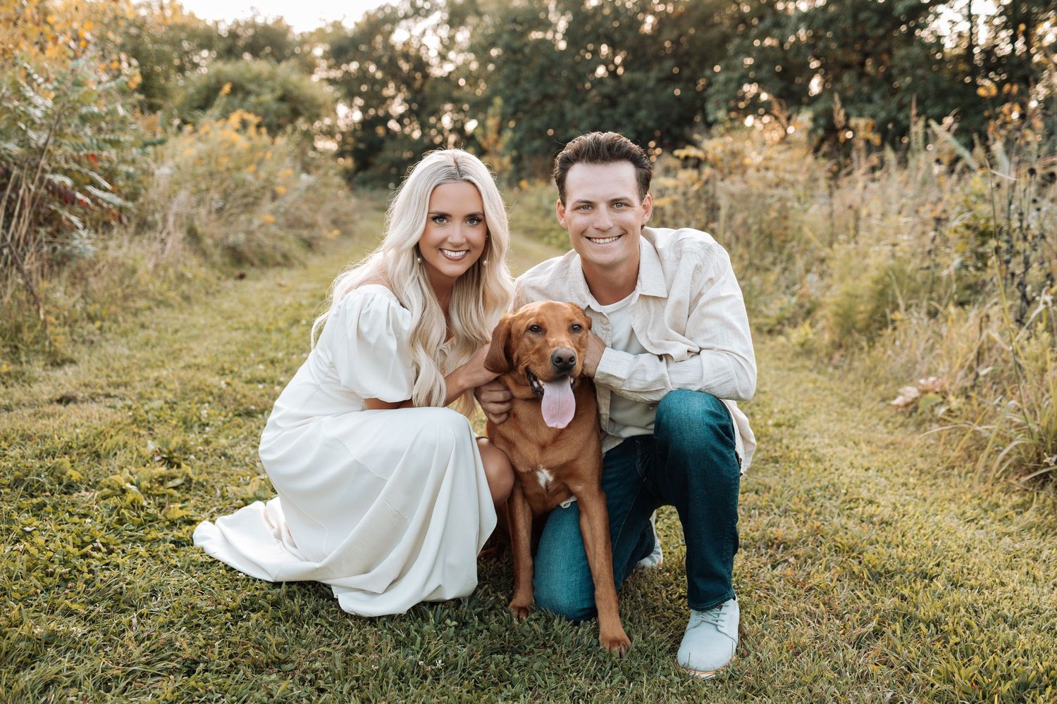Couple posing with their golden retriever dog in a sunny field during golden hour.