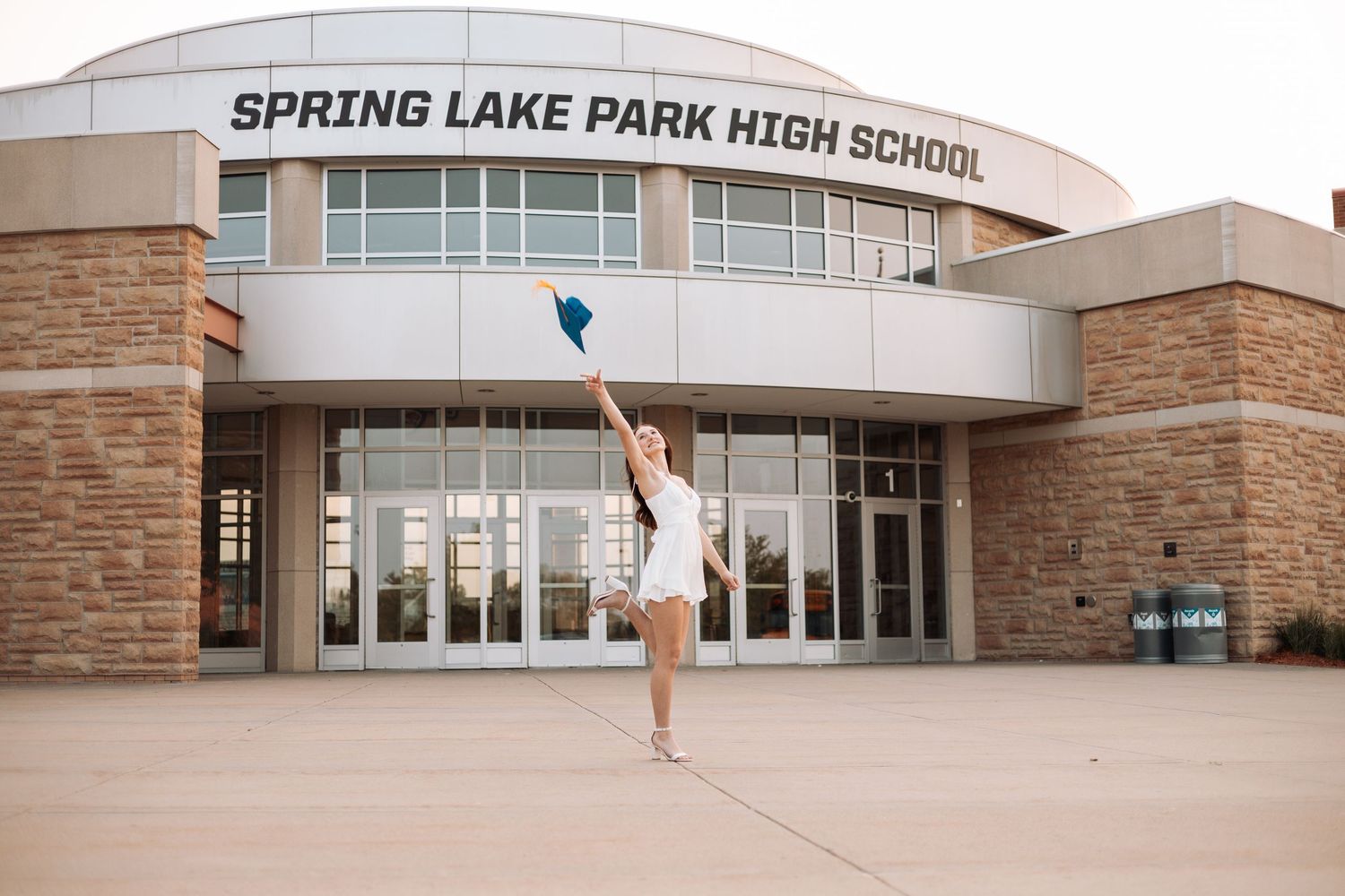 Graduate in white dress throws graduation cap in front of school building with Kenzie Ayla Images in the Twin Cities MN area.