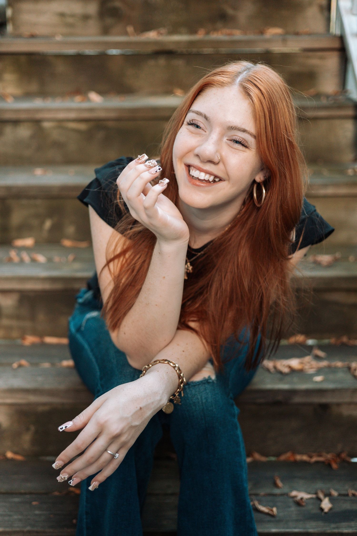 A person with red hair wearing a teal sweater laughs joyfully while sitting on wooden steps covered in autumn leaves.