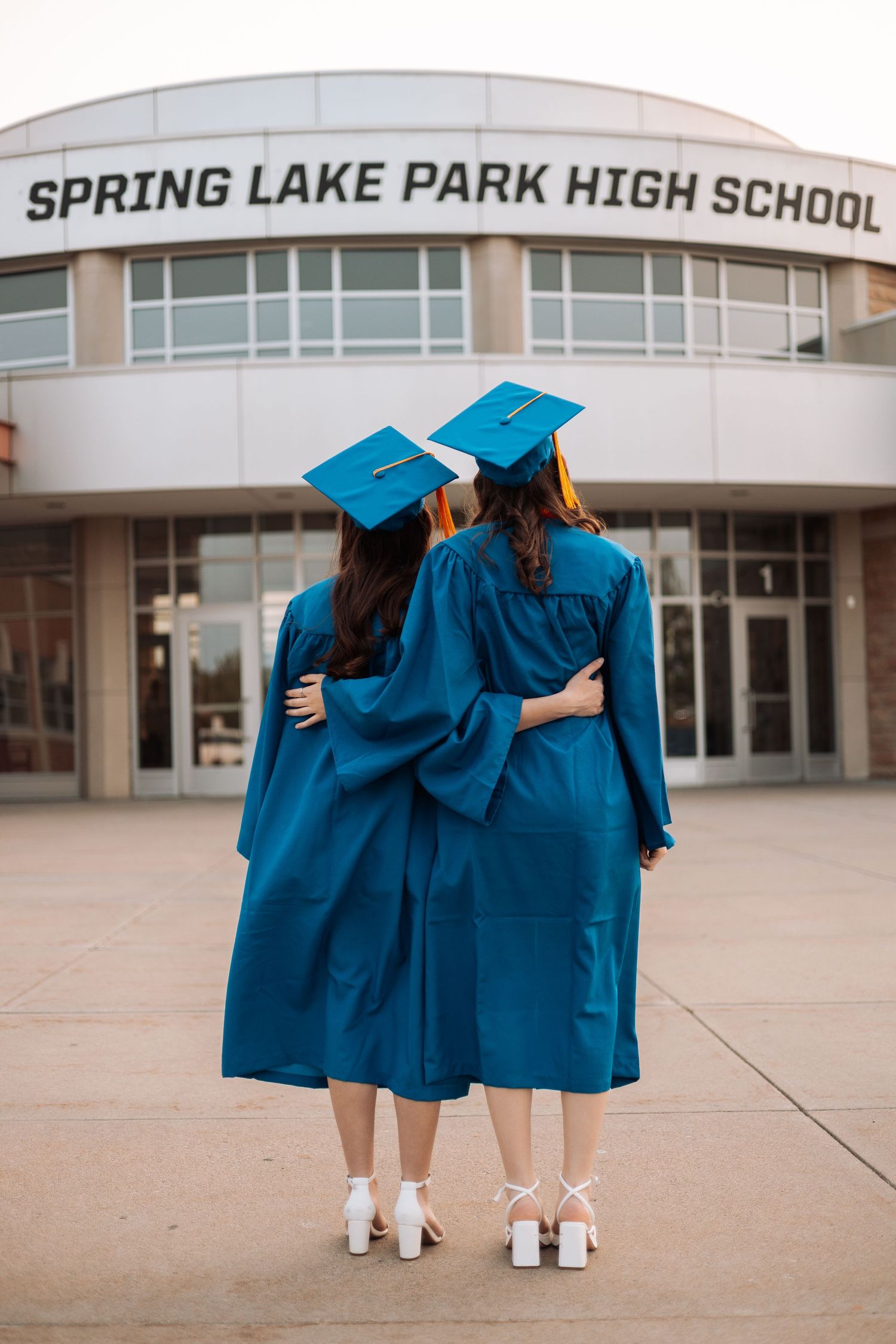 Two graduates in blue gowns embrace while facing school entrance with Kenzie Ayla Images in the Twin Cities MN area.