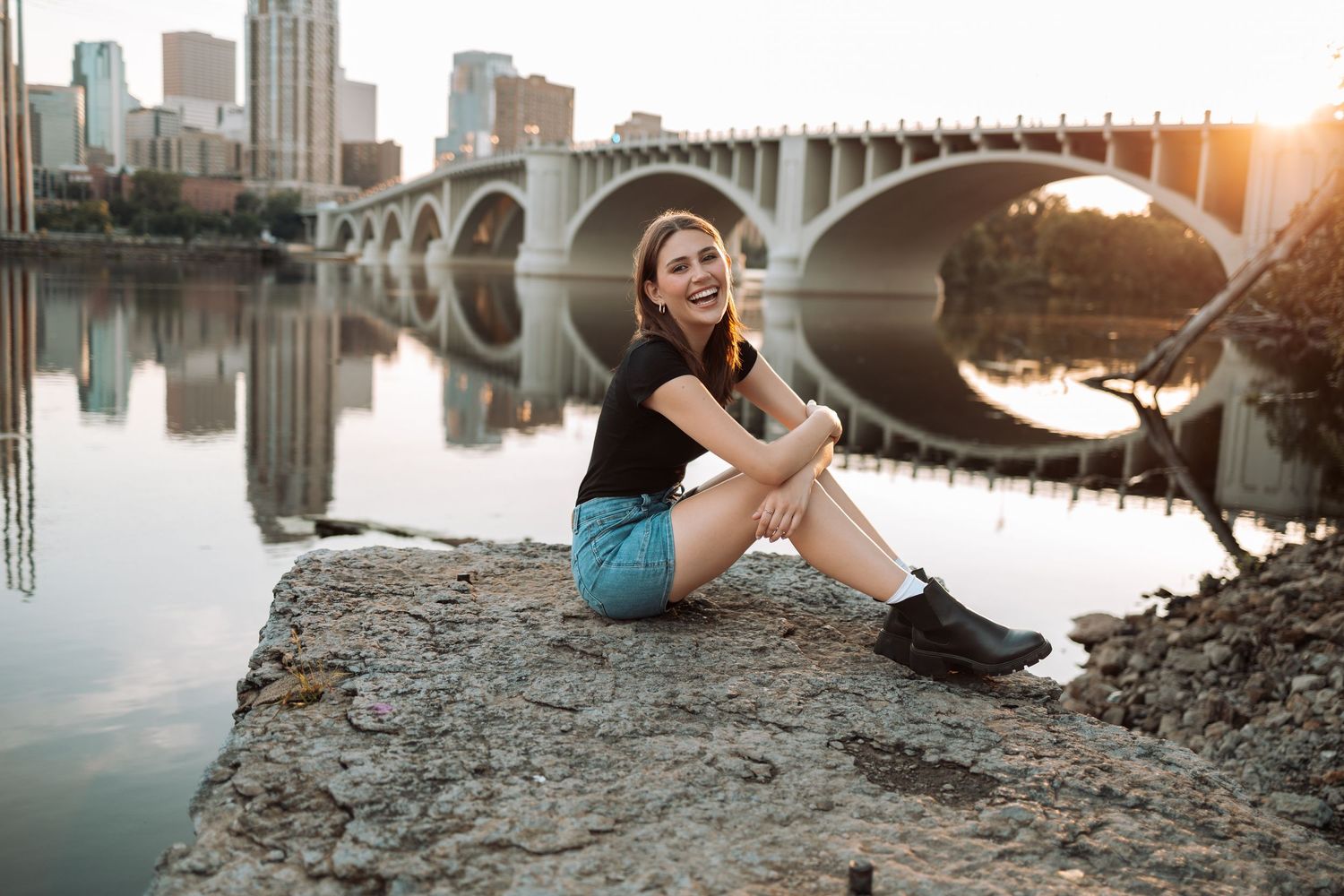 Urban senior portrait session along riverbank with historic stone arch bridge at sunset.