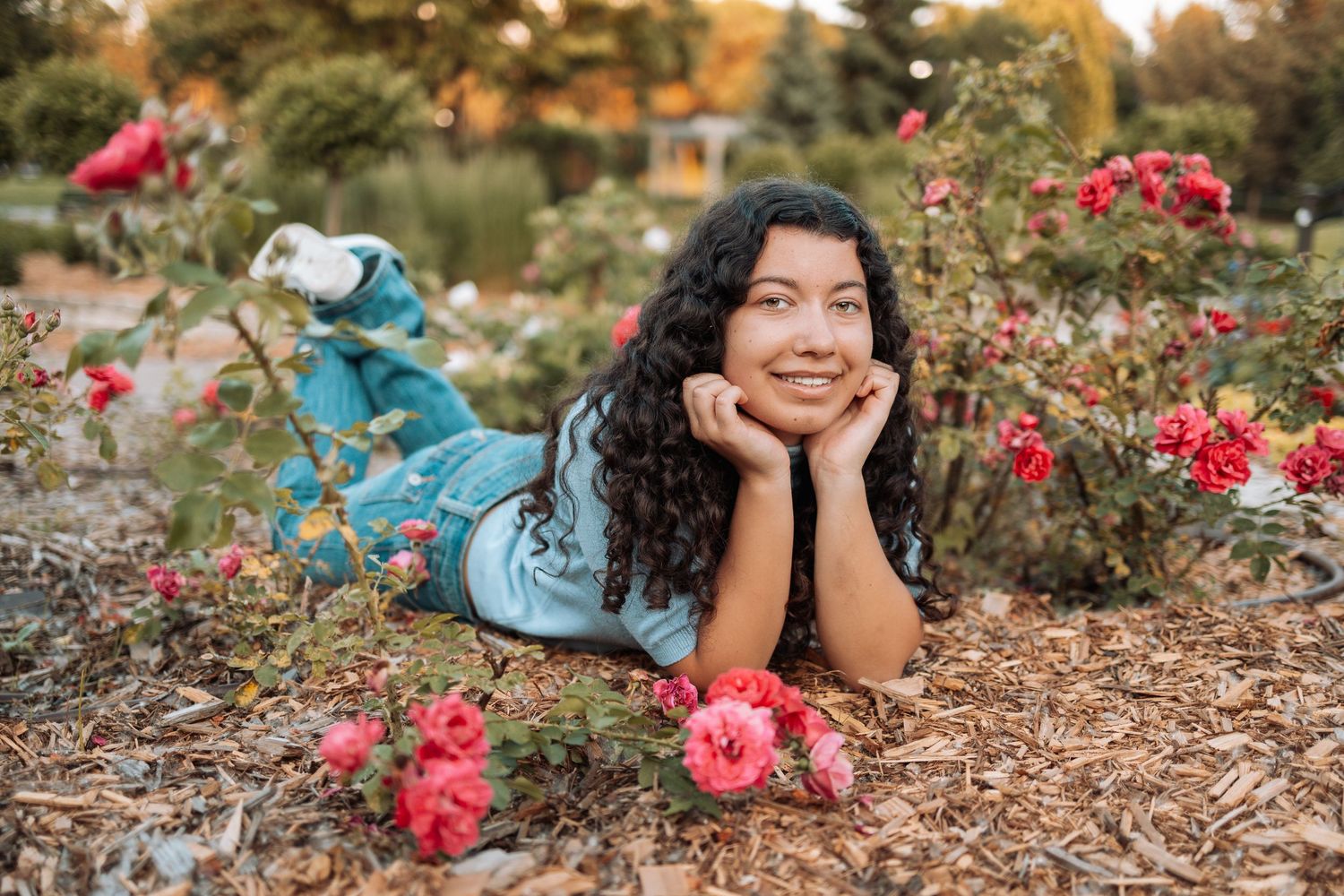 Casual poses on ground surrounded by pink roses wearing blue top and jeans with Kenzie Ayla Images in the Twin Cities MN area.