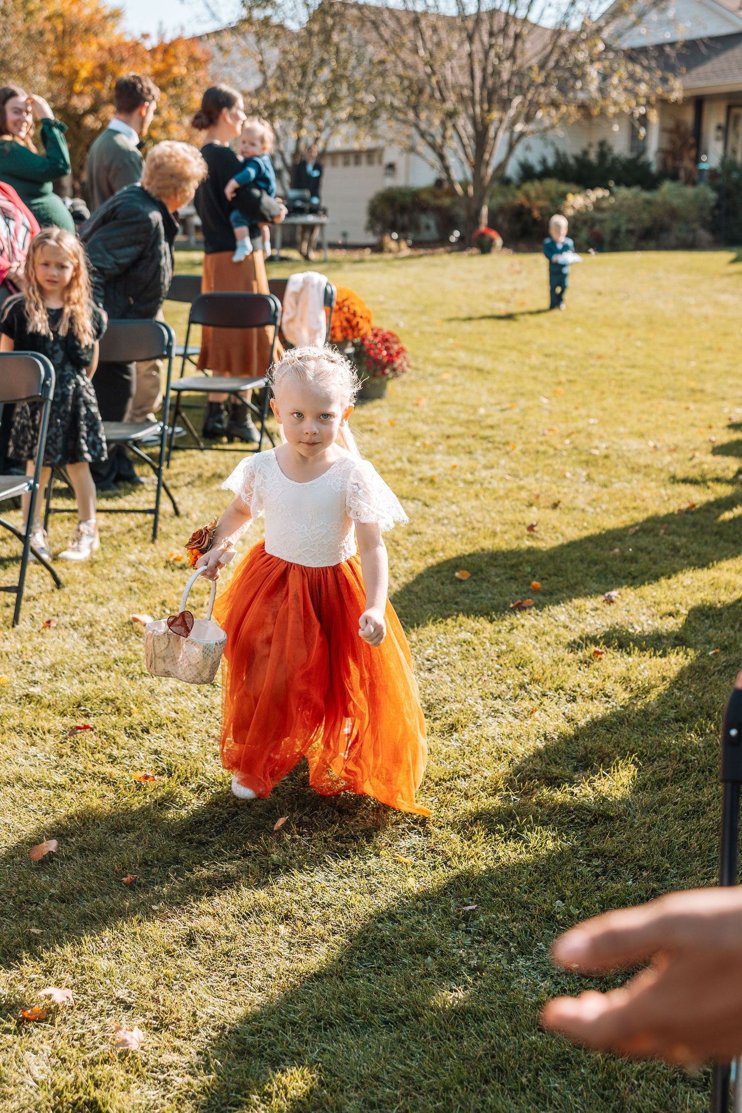 Young flower girl in white and orange tulle dress walking on grass at outdoor wedding.
