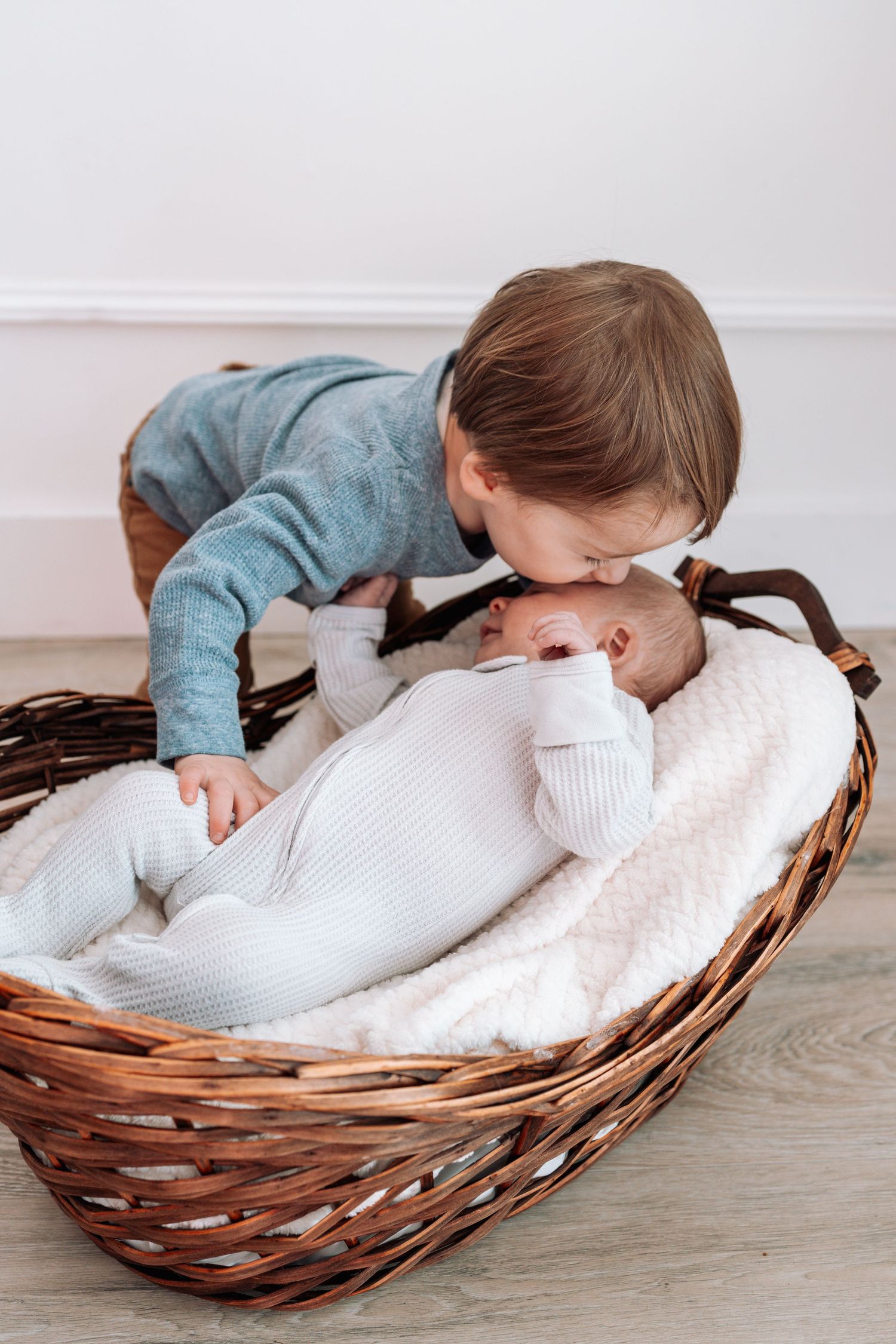 Small child peeks into woven basket with white blanket on wooden floor.