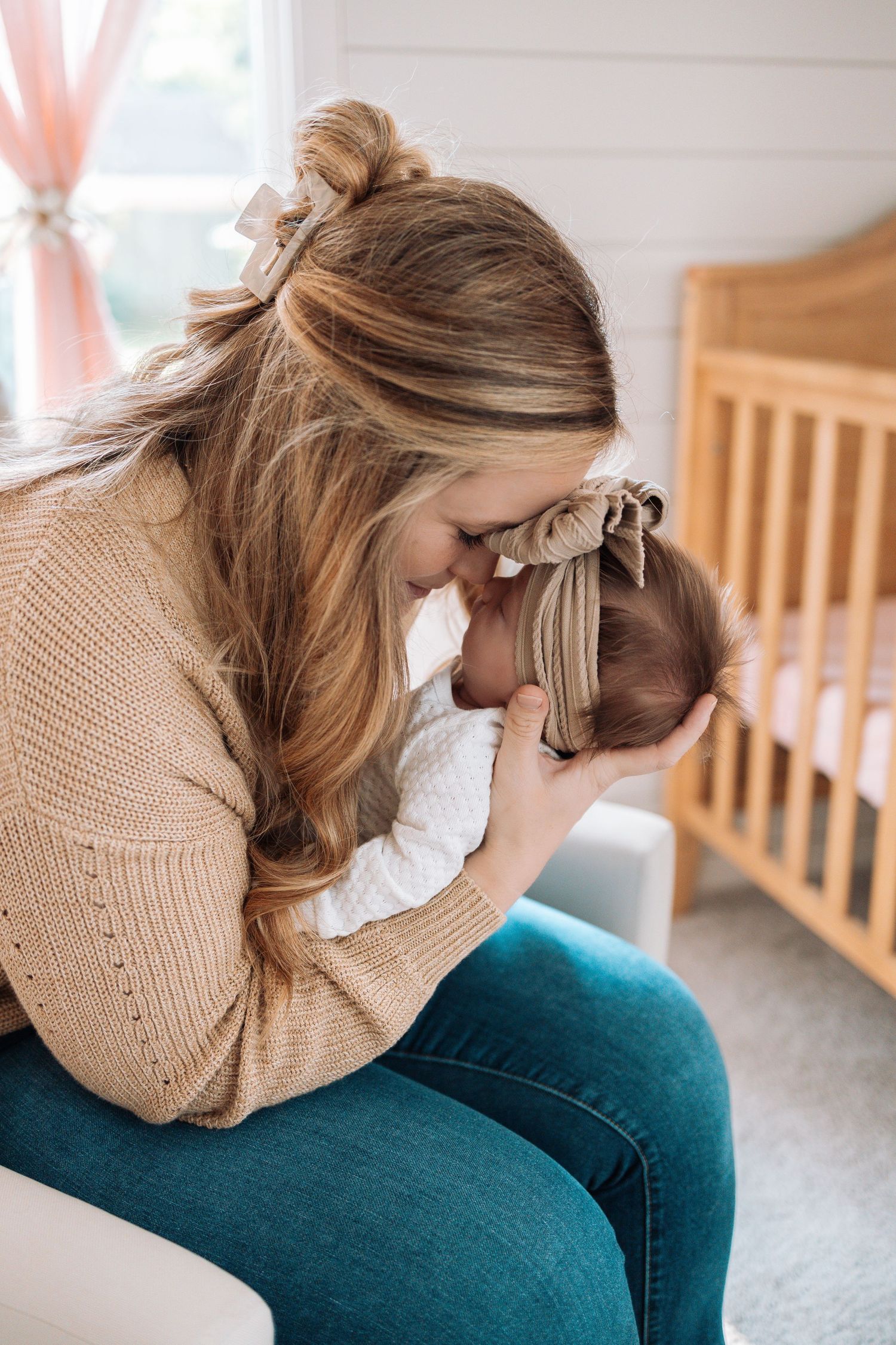 A mother in a beige sweater shares intimate moments with her baby in a nursery setting.