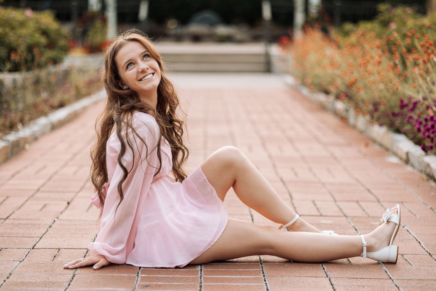 A person in a pink dress poses gracefully on a stone pathway surrounded by autumn foliage and flowers.