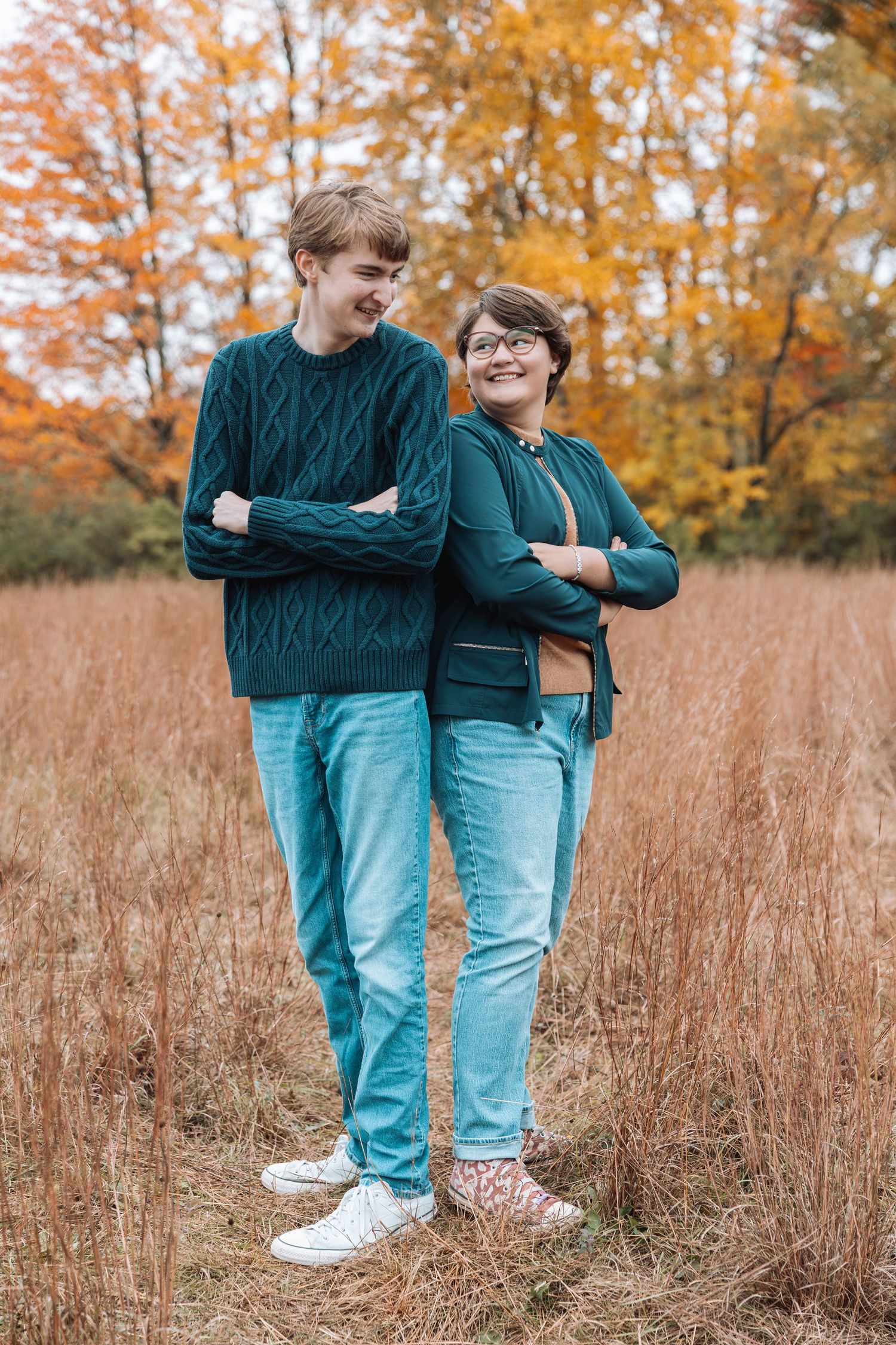 Two people in matching sweaters and jeans standing in tall autumn grass.