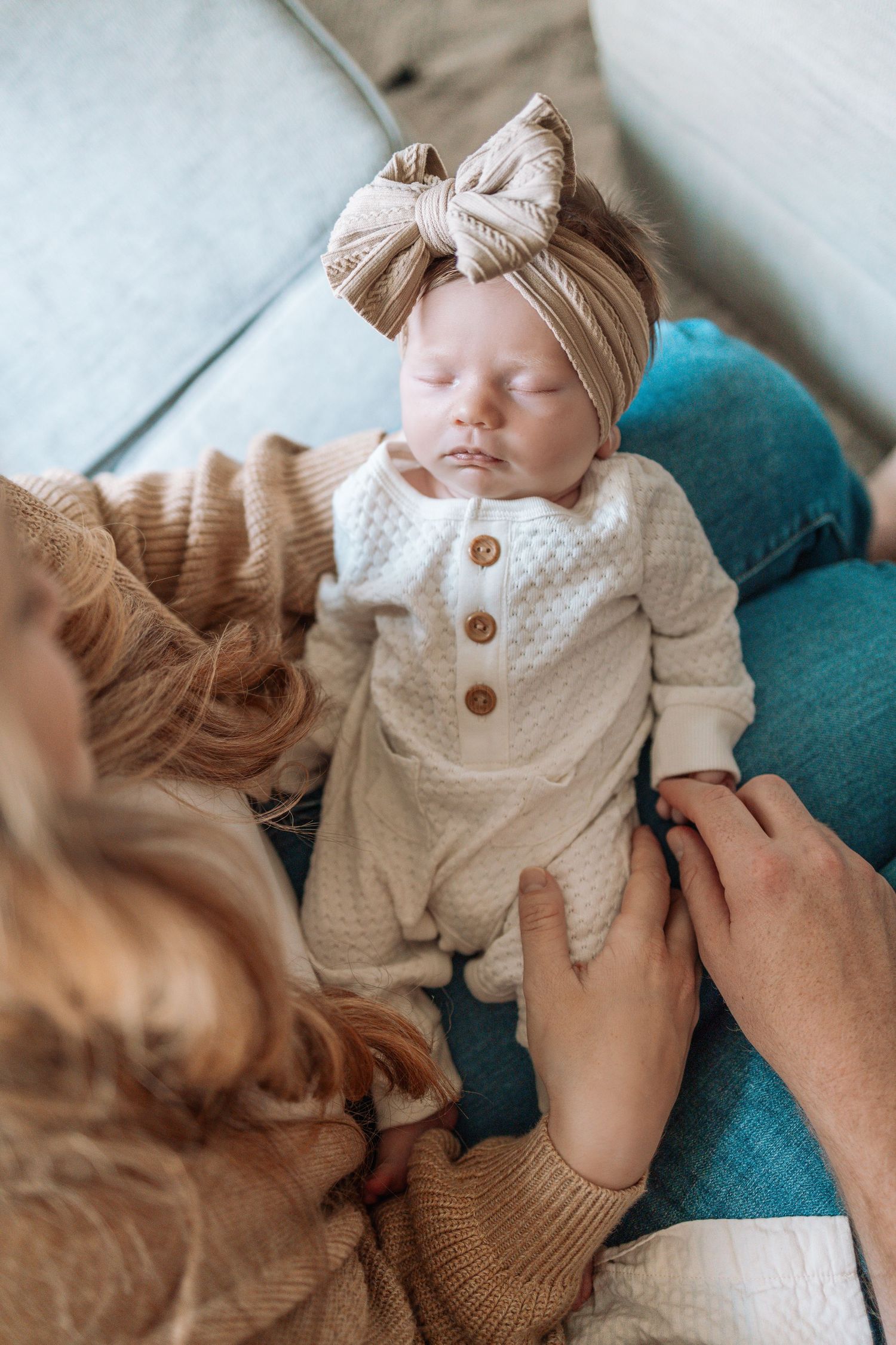 A sleeping newborn wearing a knit outfit and bow headband rests on a teal chair.