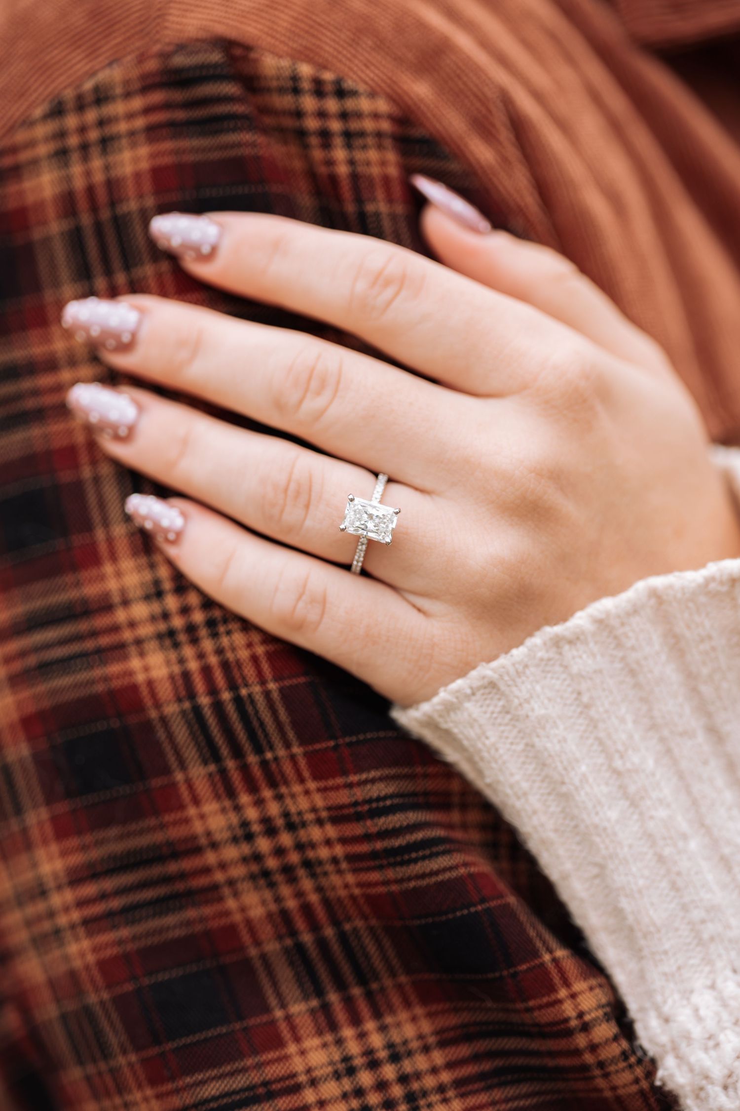 Sparkling diamond engagement ring displayed on hand against plaid flannel background.