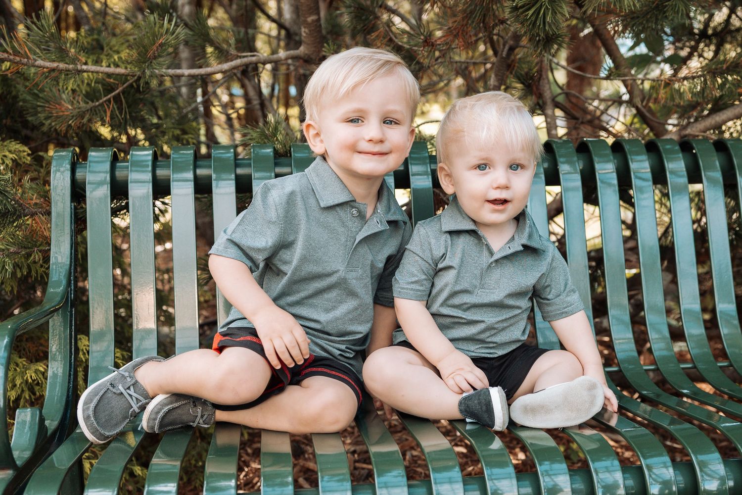Two children in matching grey shirts sitting on a green park bench.