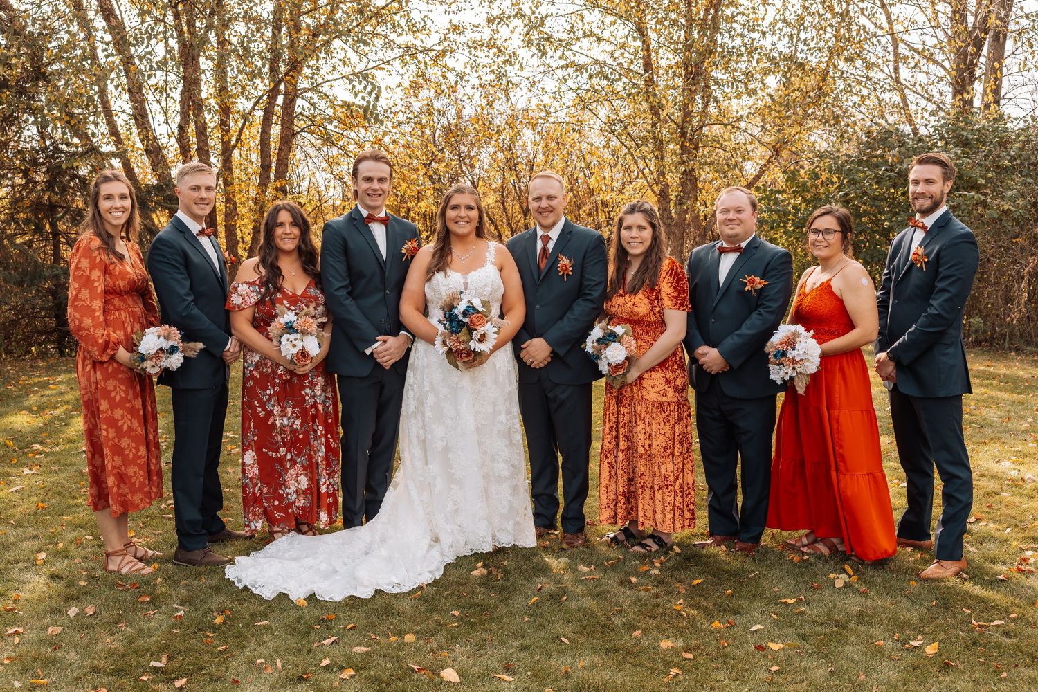 Wedding party in navy suits and burnt orange floral dresses poses together during autumn outdoor photoshoot.