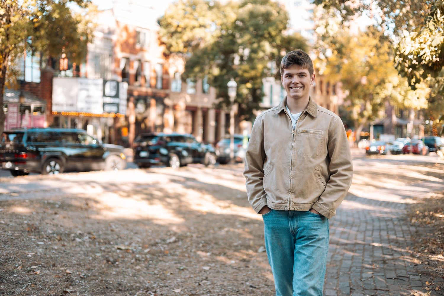 A person wearing blue jeans and a beige sweater stands in a sunlit autumn street with fallen leaves.