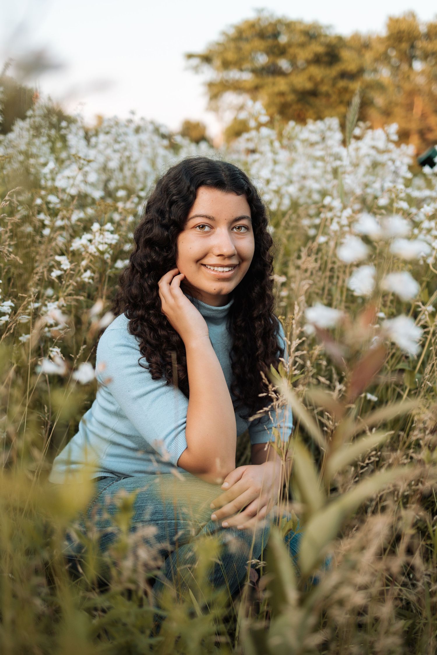 A person sits among blooming white wildflowers in a field wearing a light blue top on a sunny afternoon.