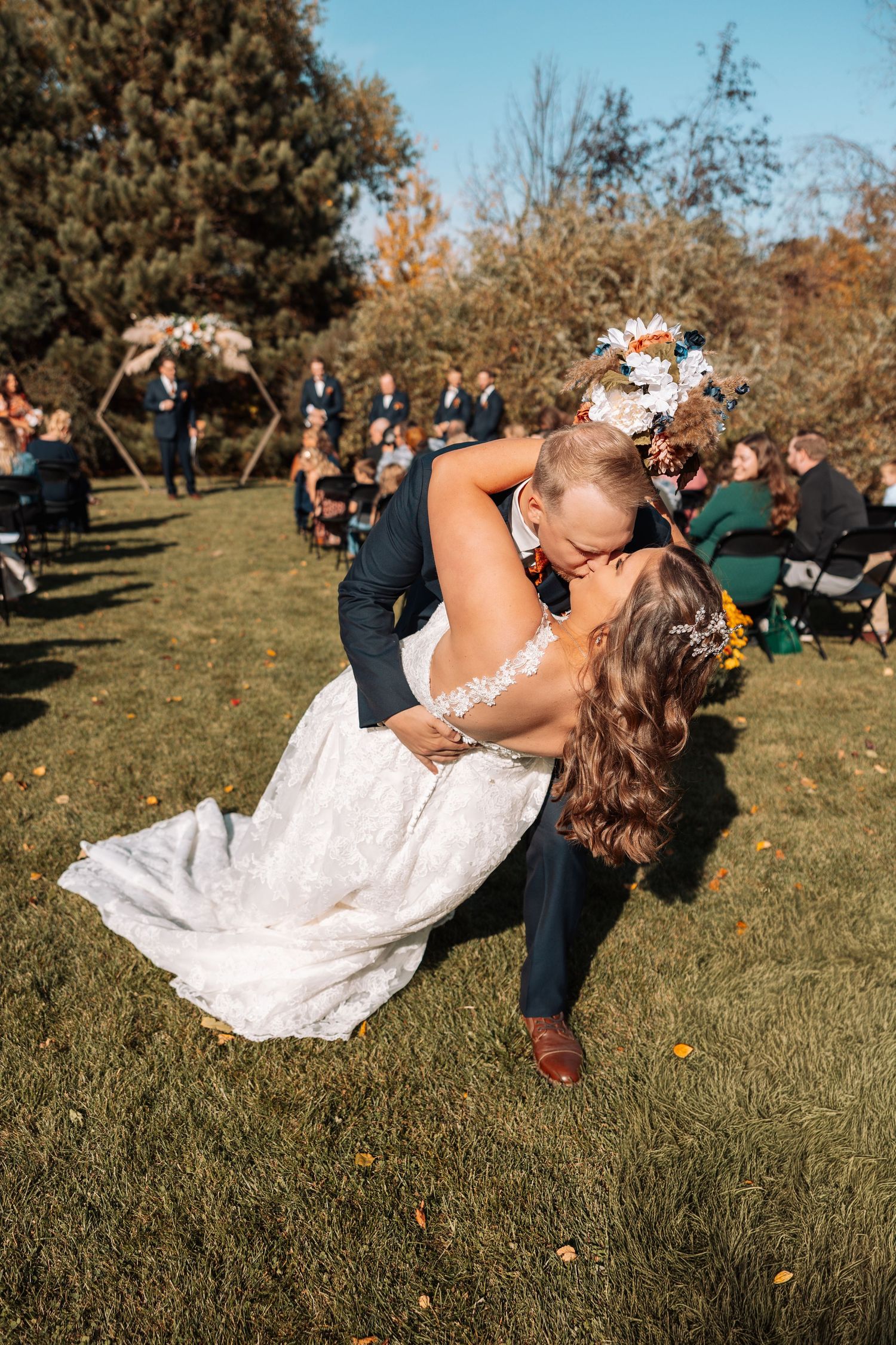 Couple in wedding attire performs dramatic dip pose on grassy field.