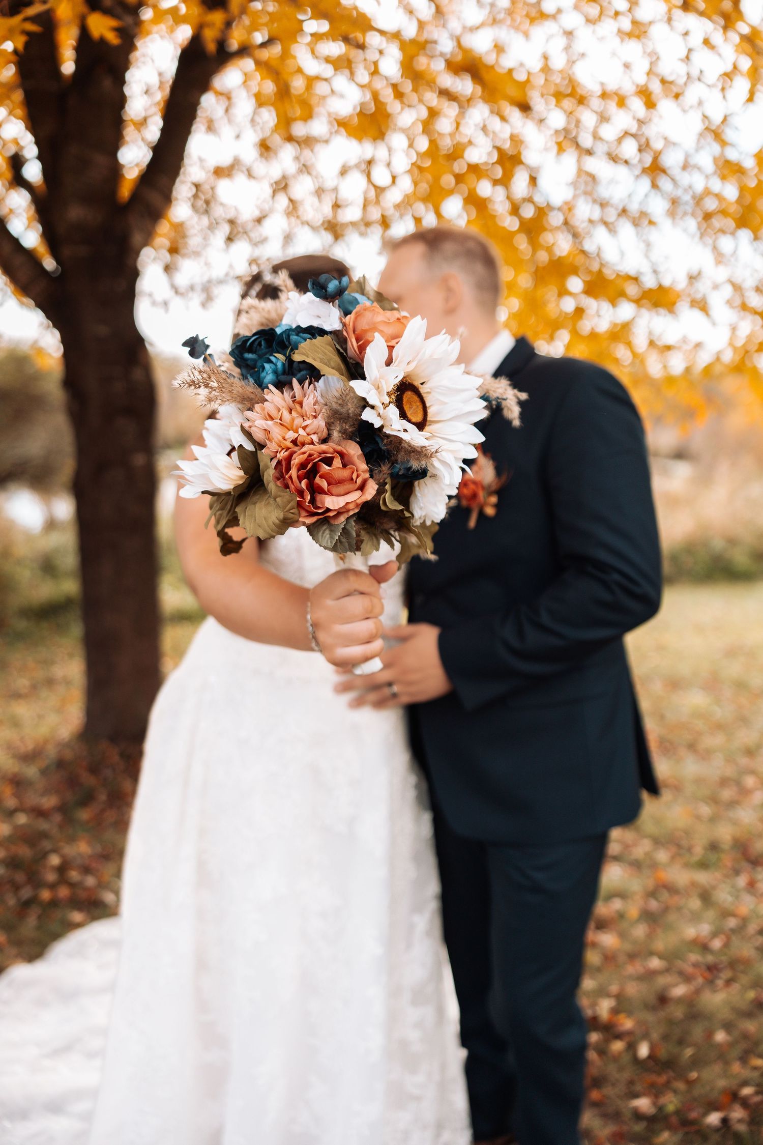 A wedding couple holding a fall-themed bouquet with orange and blue flowers against autumn foliage.