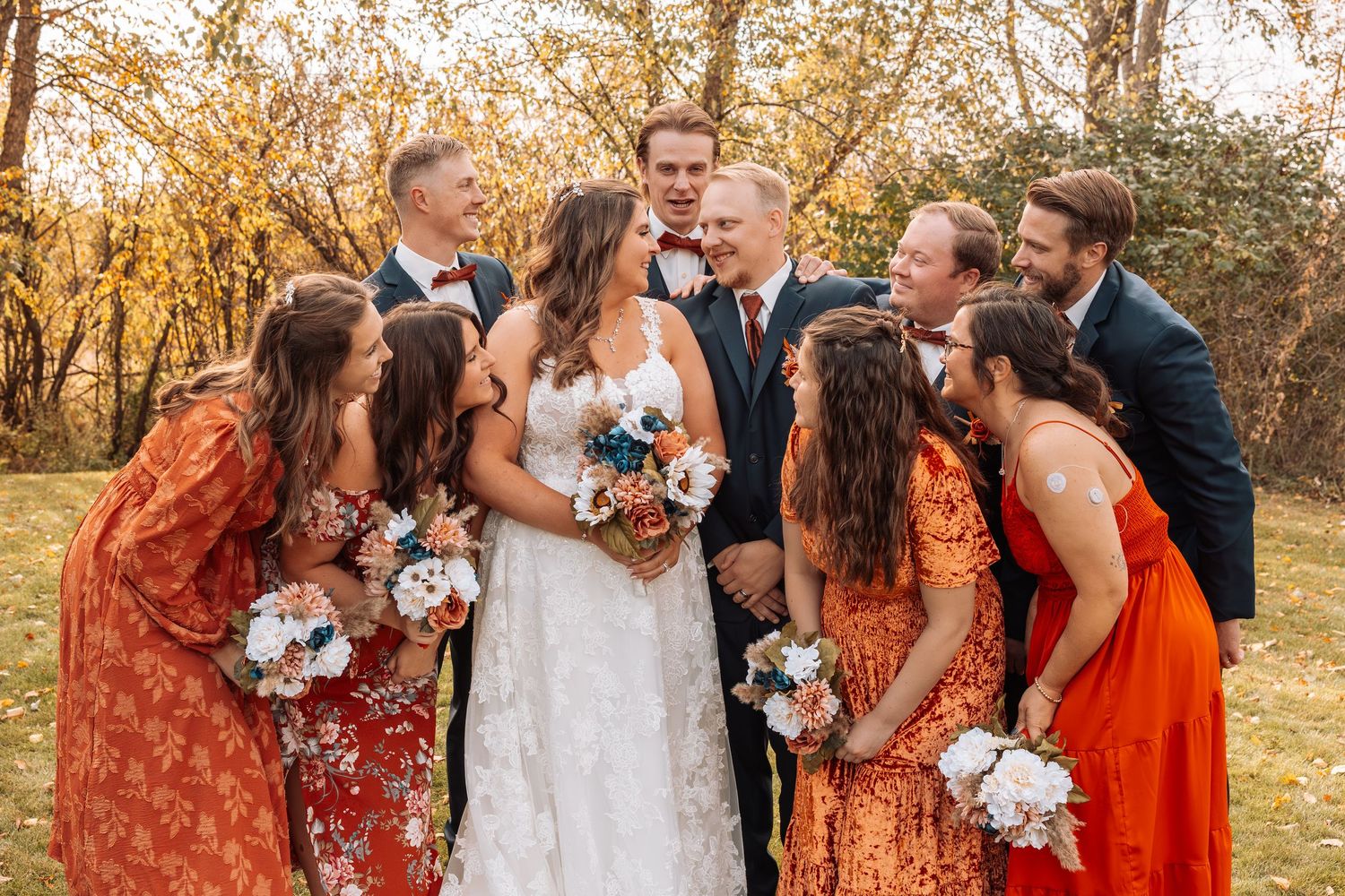 Wedding group photo with bridesmaids in orange floral dresses and groomsmen in navy suits.