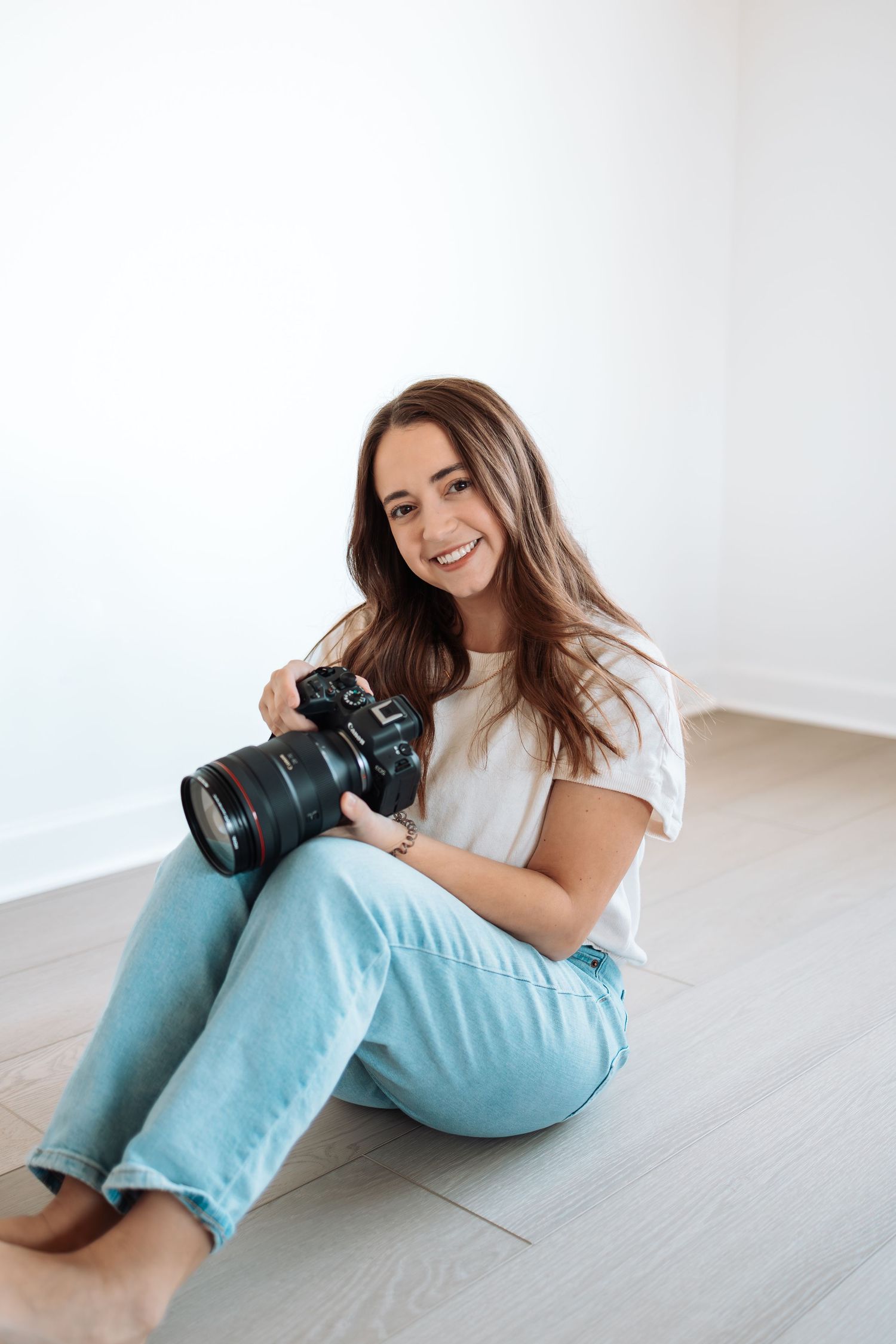 Professional photographer in light blue jeans and white top poses with camera while sitting on white floor.