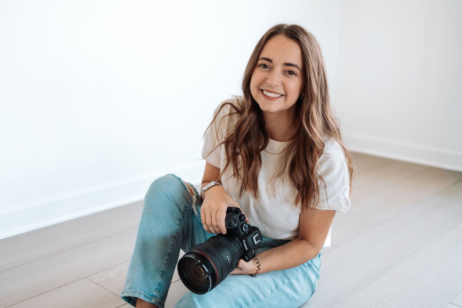 Photographer in white t-shirt and blue jeans sits on floor holding professional camera while smiling.