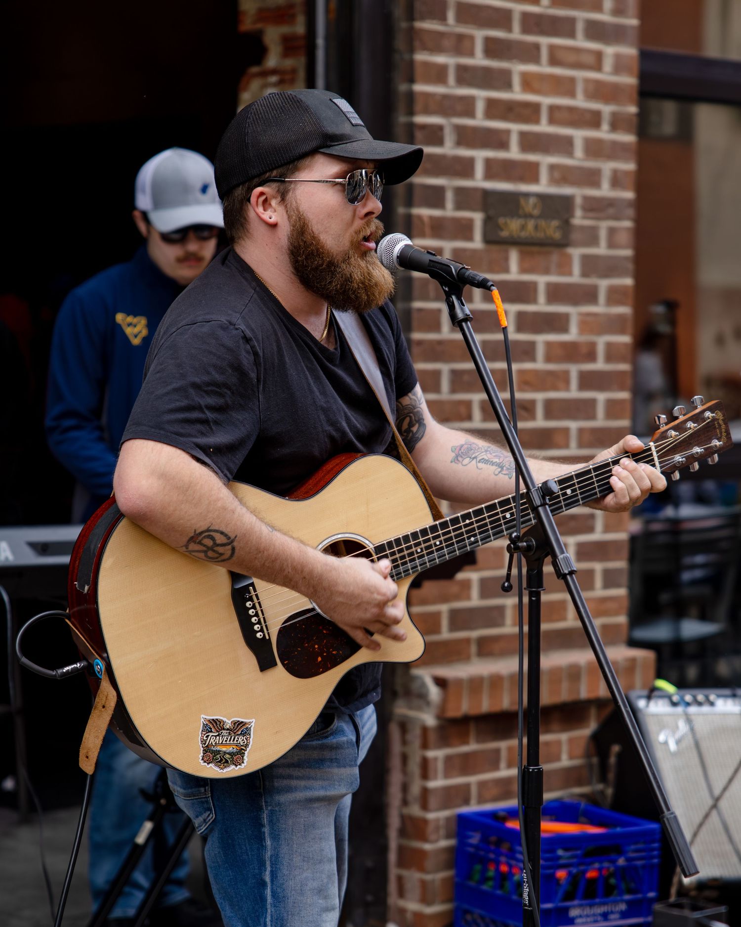 Bearded male musician playing acoustic guitar and singing into microphone outdoors near brick building wearing black cap.