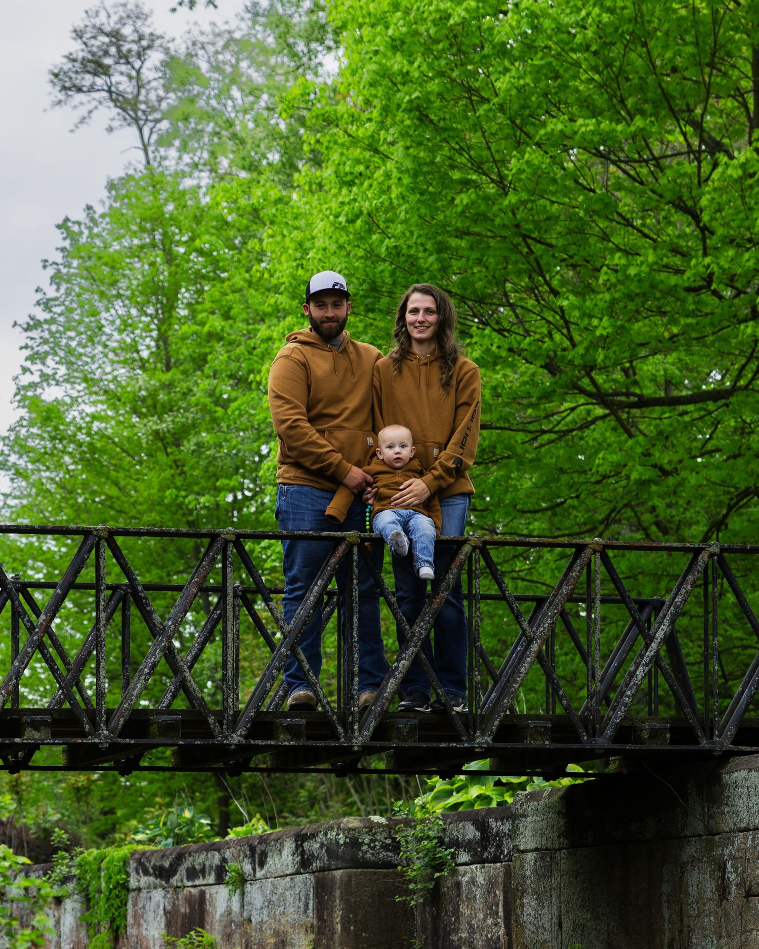 Smiling family in matching brown hoodies poses on rustic iron bridge surrounded by green trees.