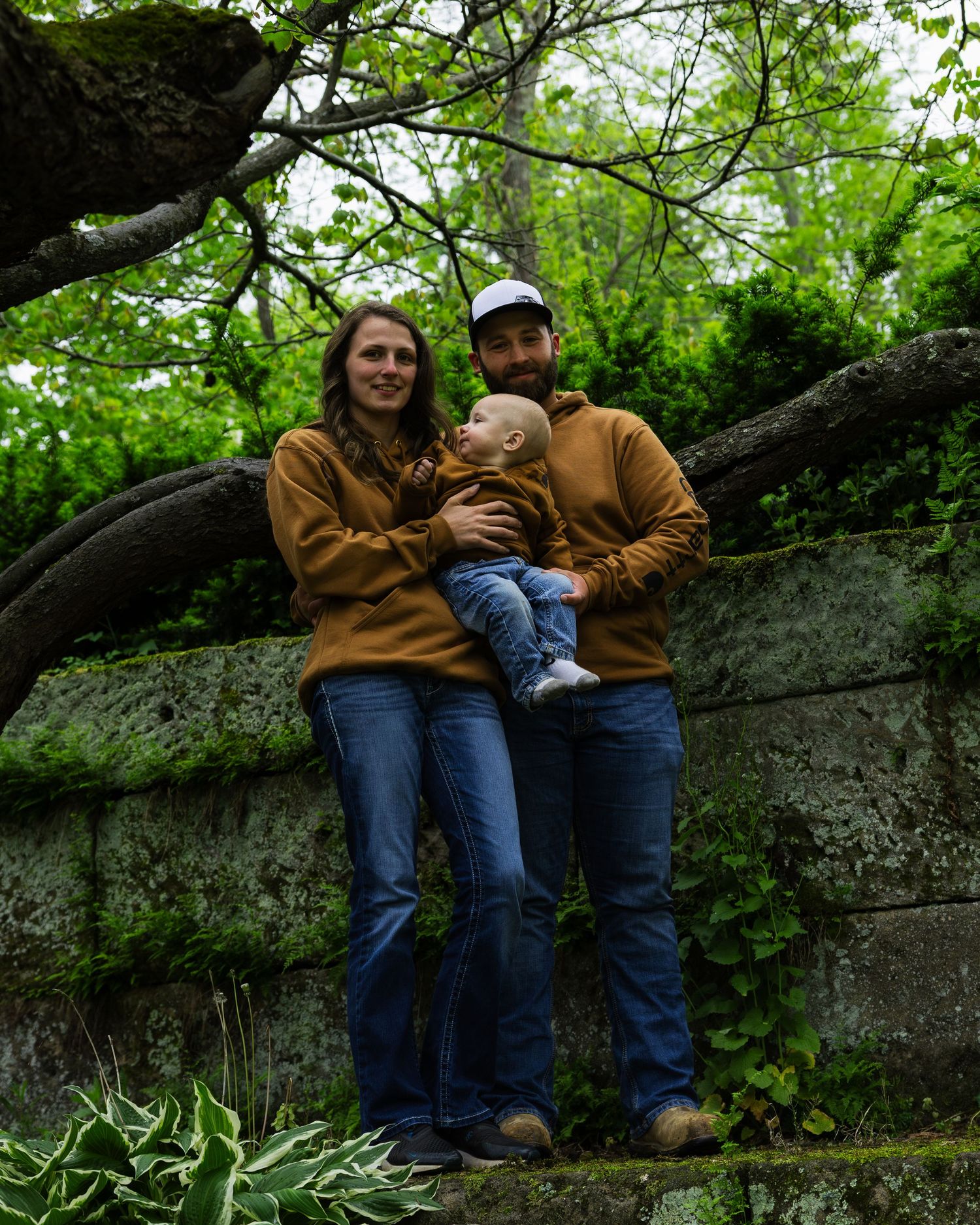 Happy couple in matching brown hoodies holds baby against mossy stone wall outdoors.
