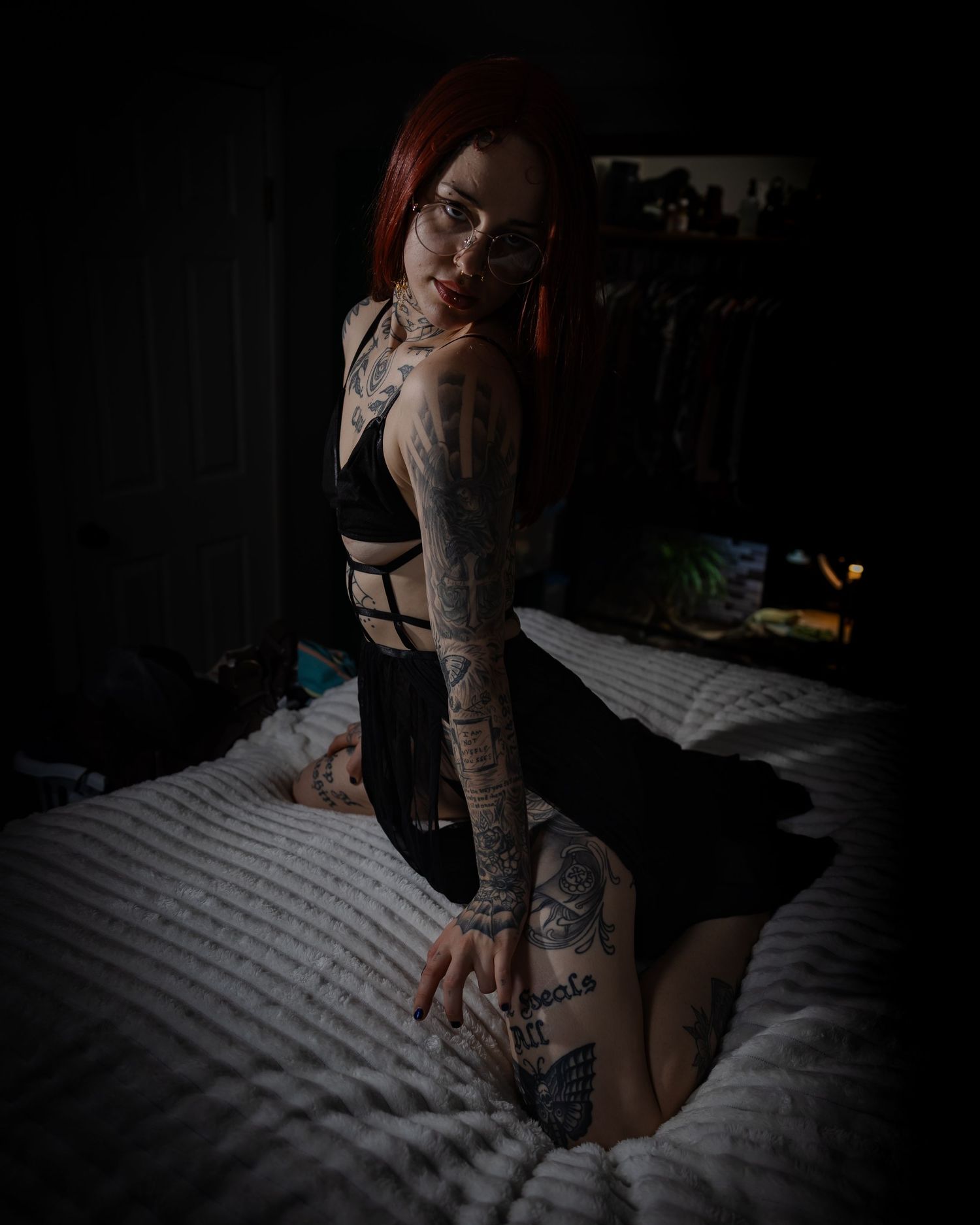 A tattooed redhead woman in black lingerie poses on a bed in a dimly lit bedroom.