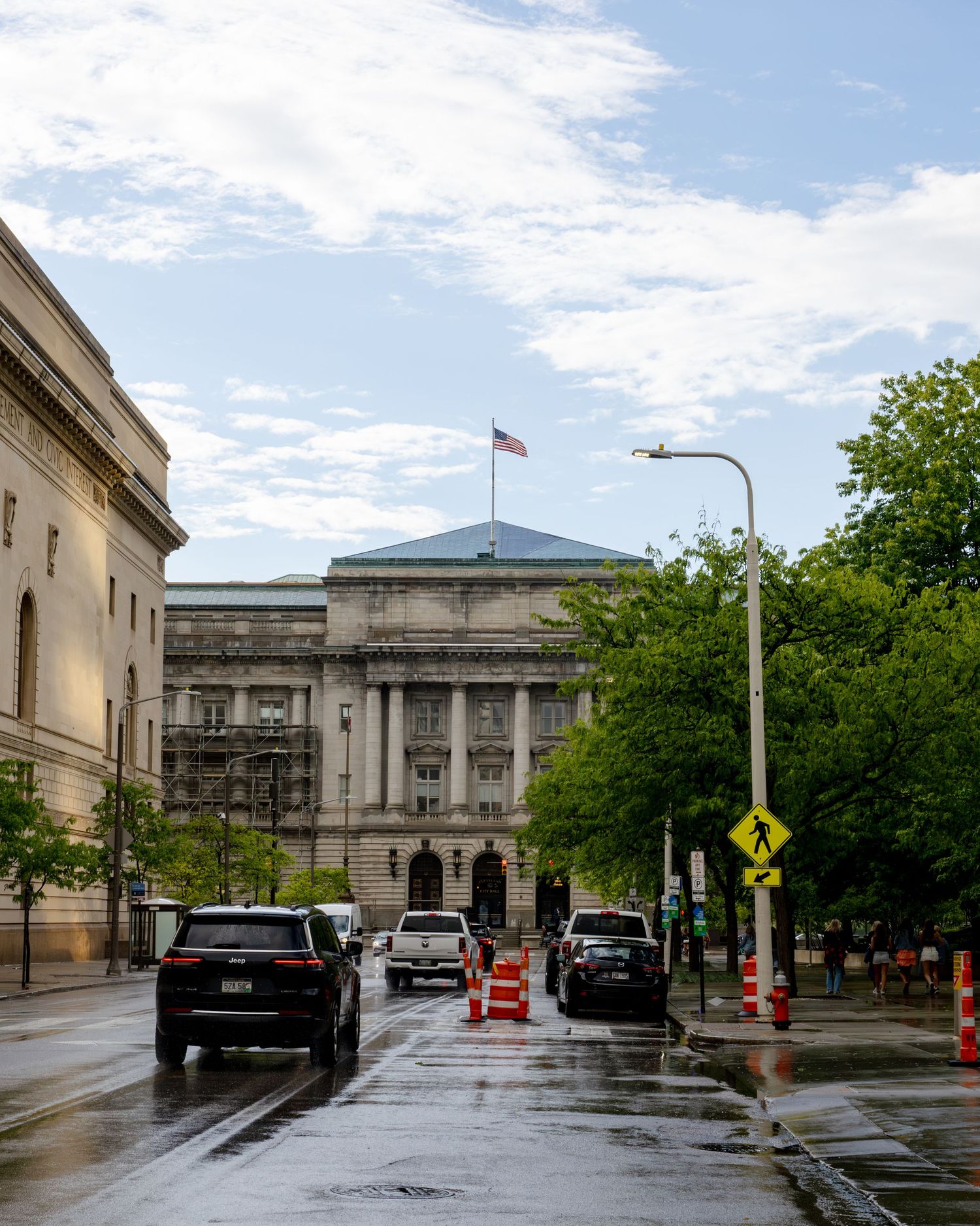Rainy day street view of a neoclassical government building with American flag, wet roads, and green trees in Washington DC.