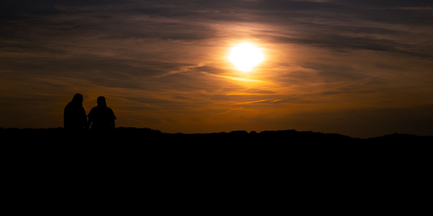 Two silhouetted figures sit together watching a vibrant golden sunset over a dark horizon.