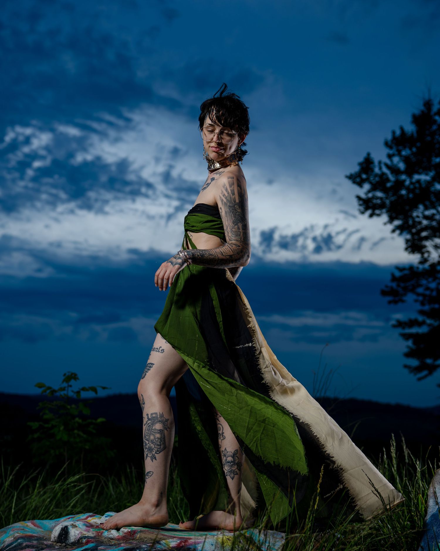 Tattooed woman in green draped outfit poses outdoors at twilight with moody blue sky.